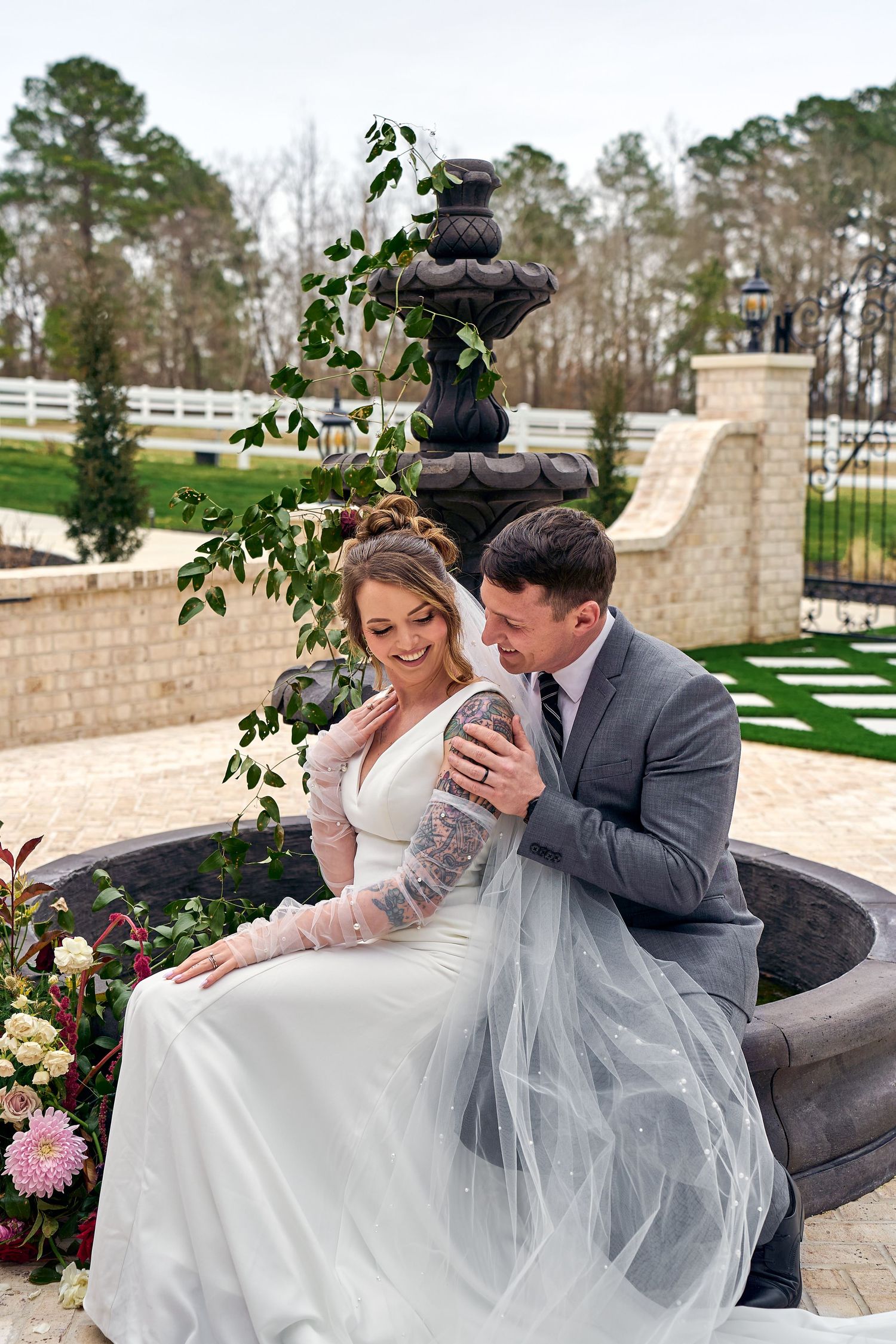 Newlyweds share a tender moment sitting by an elegant stone fountain in a garden setting with white fencing and pine trees.