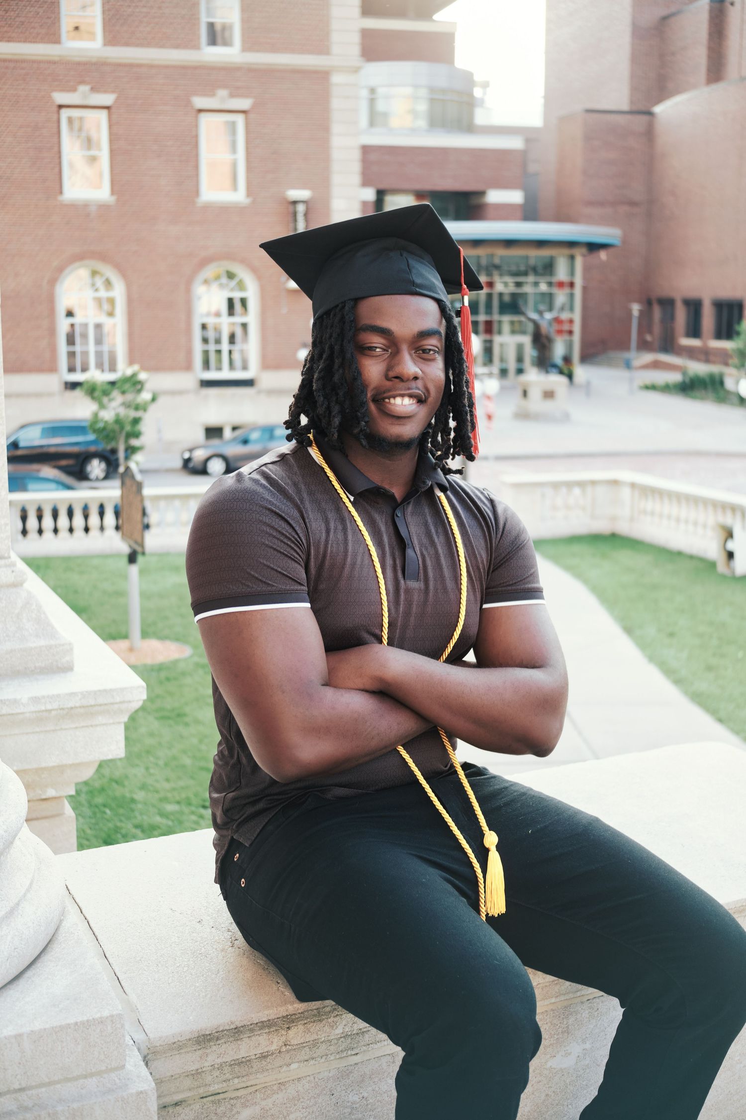 Smiling graduate in cap and gold stole sitting with arms crossed on campus steps.