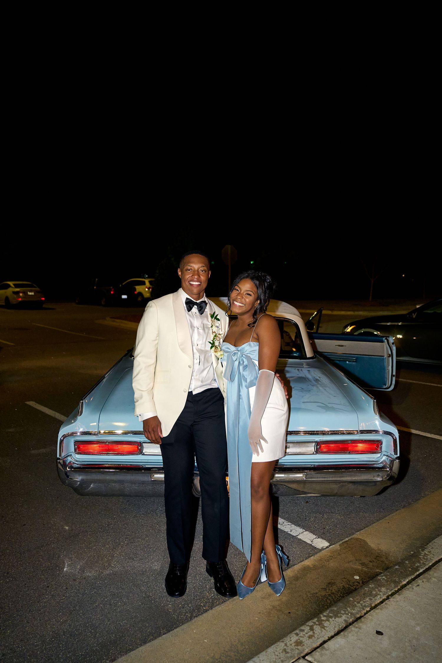Newlyweds pose beside a classic light blue vintage car at night after their wedding reception.