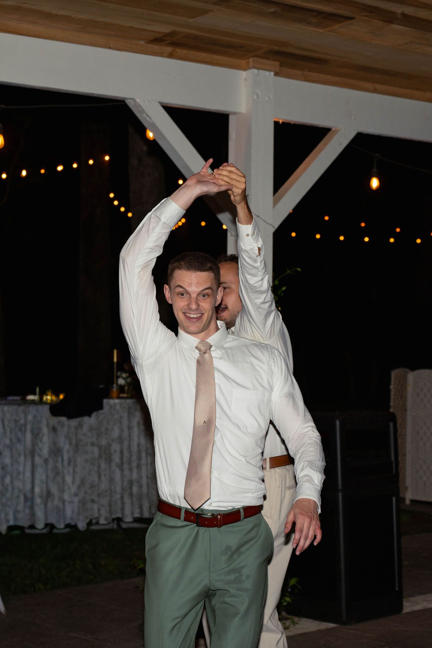 Groomsman in white shirt and tan tie dancing with raised arms at a wedding reception.
