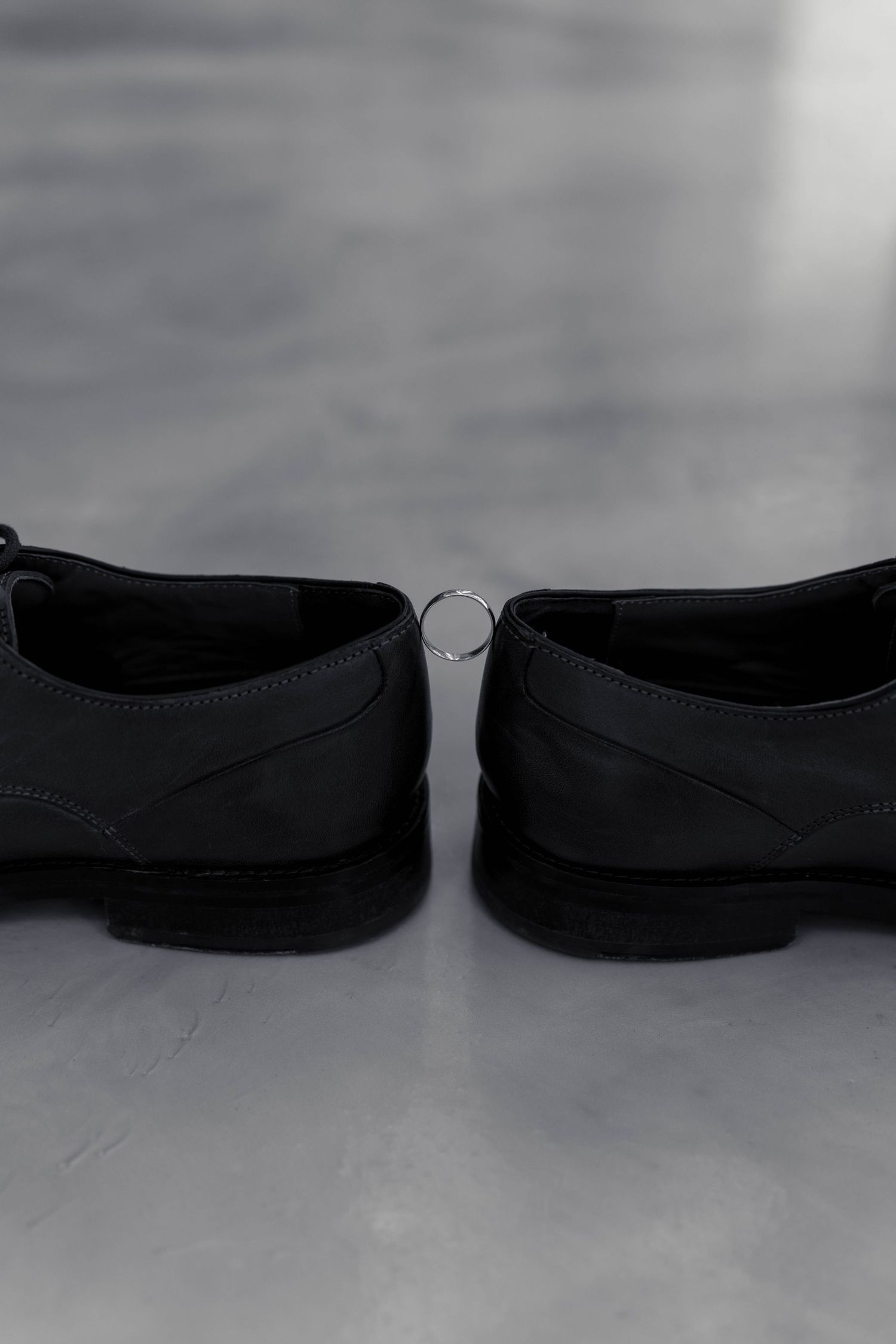 Two black dress shoes with a silver wedding ring placed between them on gray surface.
