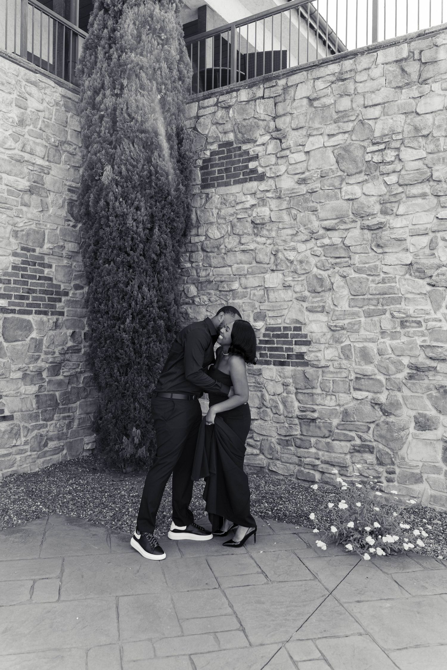 Black and white photo of couple sharing intimate kiss against stone wall with cypress tree backdrop.