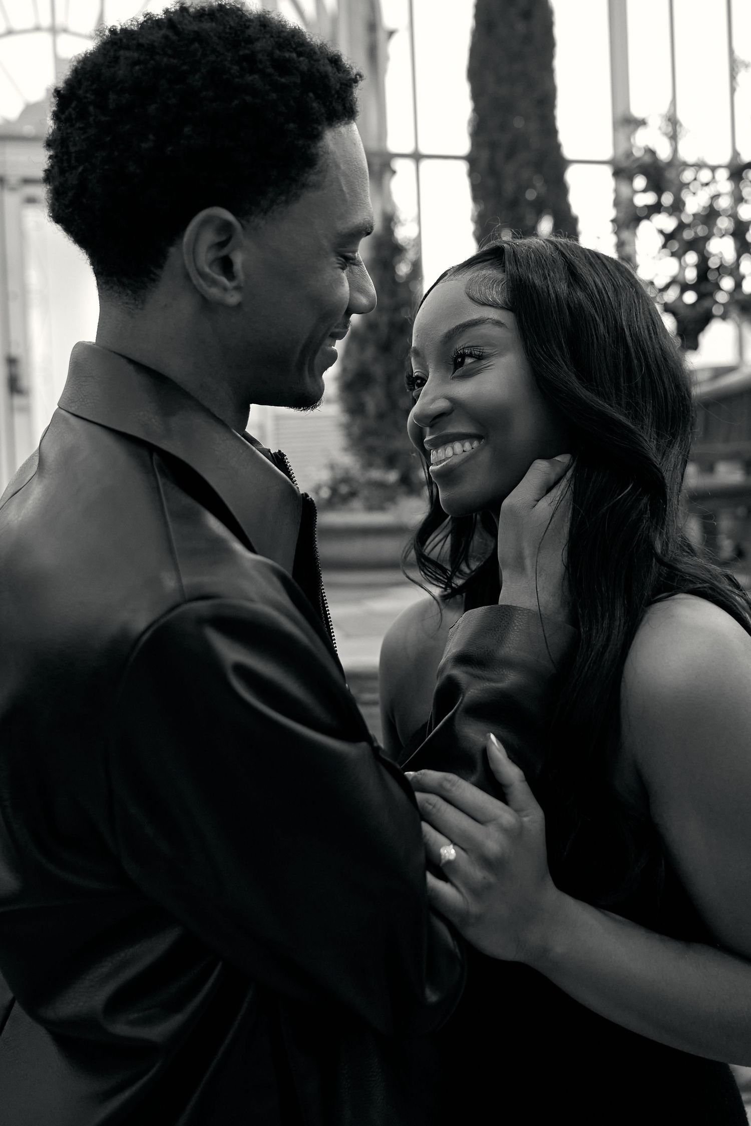 Black and white romantic photo of smiling engaged couple facing each other inside a beautiful glass conservatory.