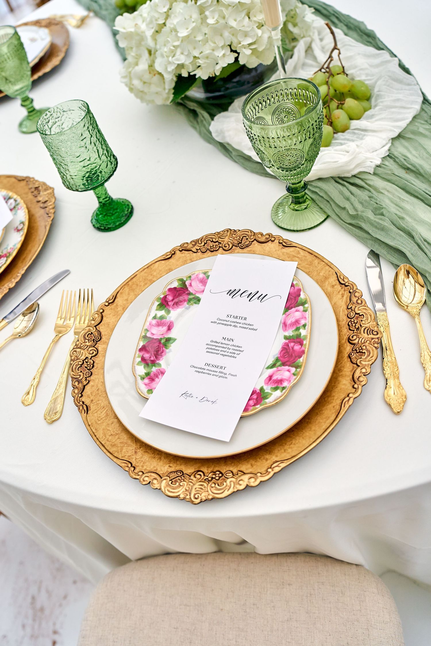 Elegant table setting with ornate gold charger plate, floral menu card, green vintage glasses and white hydrangeas.