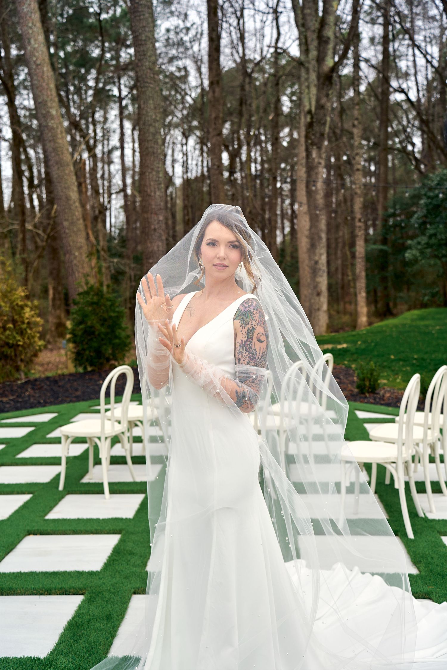 A beautiful wedding ceremony setup in a wooded outdoor venue with white chairs and stone pathway on artificial grass.