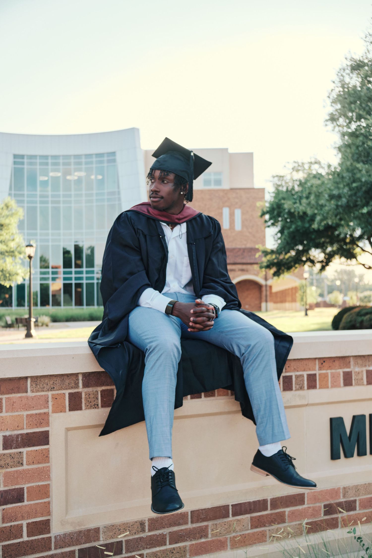 Male graduate in cap and gown sitting on a brick wall on a university campus.