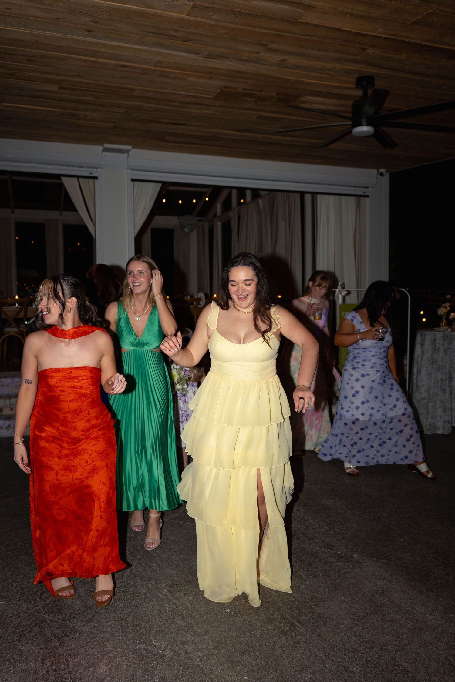 Wedding guests in colorful formal gowns dancing joyfully at an evening wedding reception.