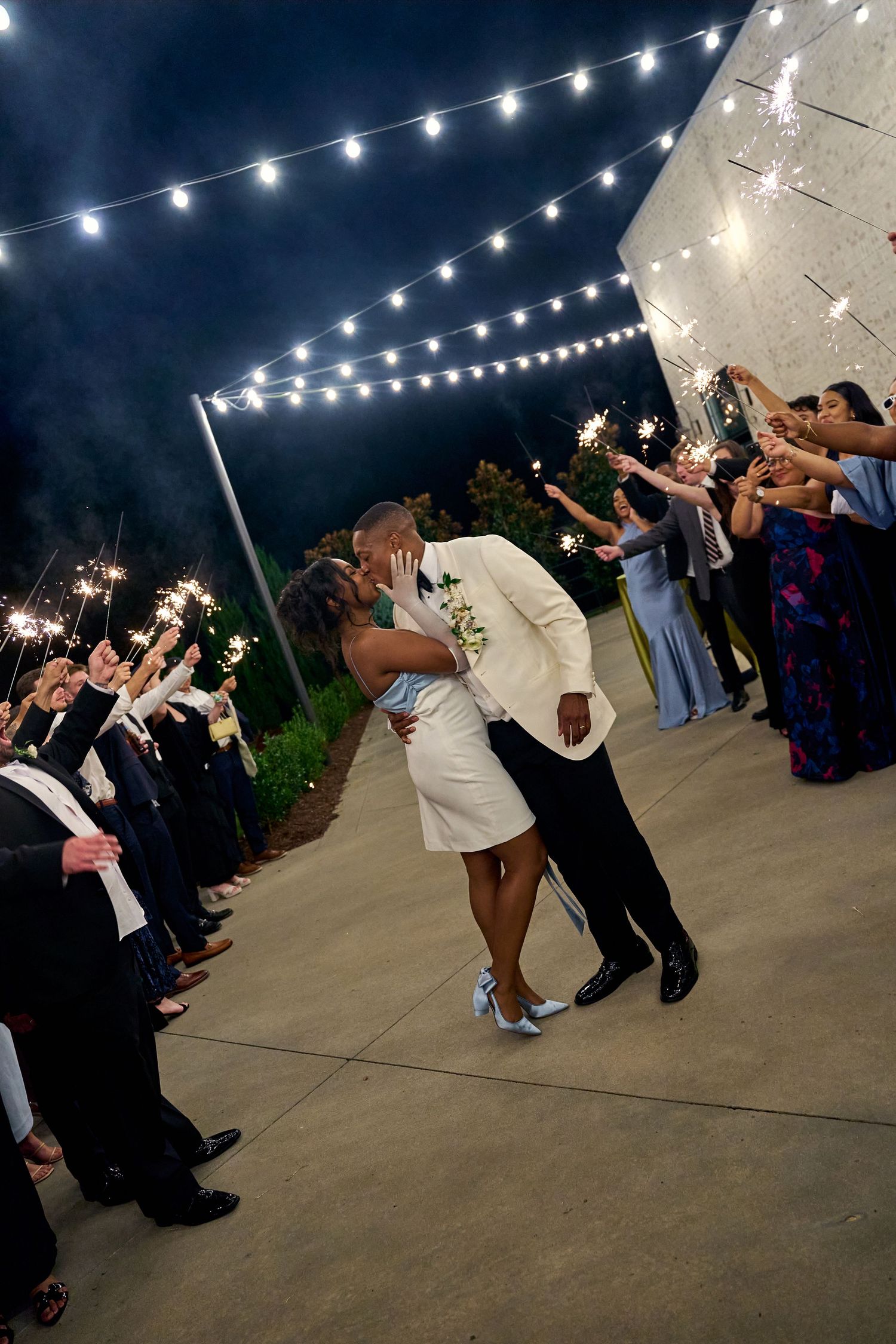 Bride and groom share a romantic kiss during their sparkler exit under twinkling string lights.