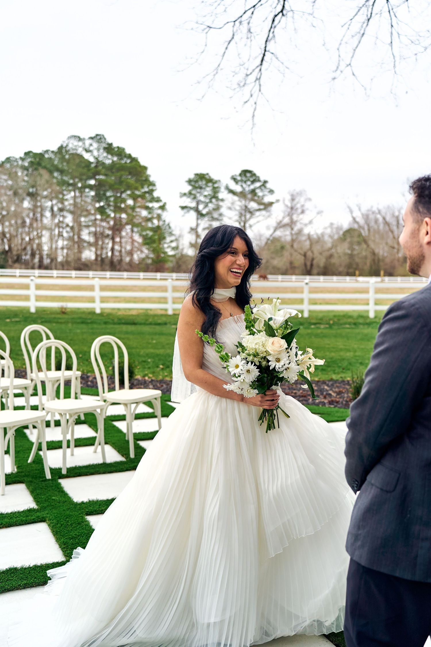 A bride in a flowing white pleated gown holds a bouquet at an outdoor garden ceremony with white chairs and fencing.