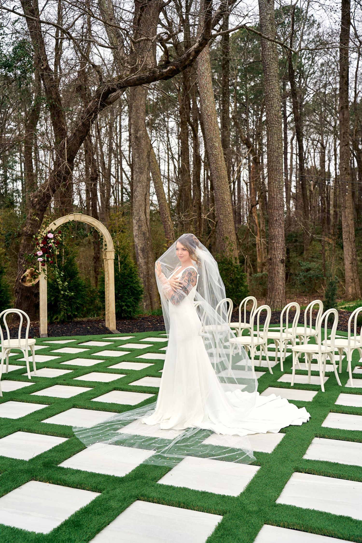 Elegant outdoor wedding venue features a wooden arch and checkerboard turf design with white chairs in a forest setting.