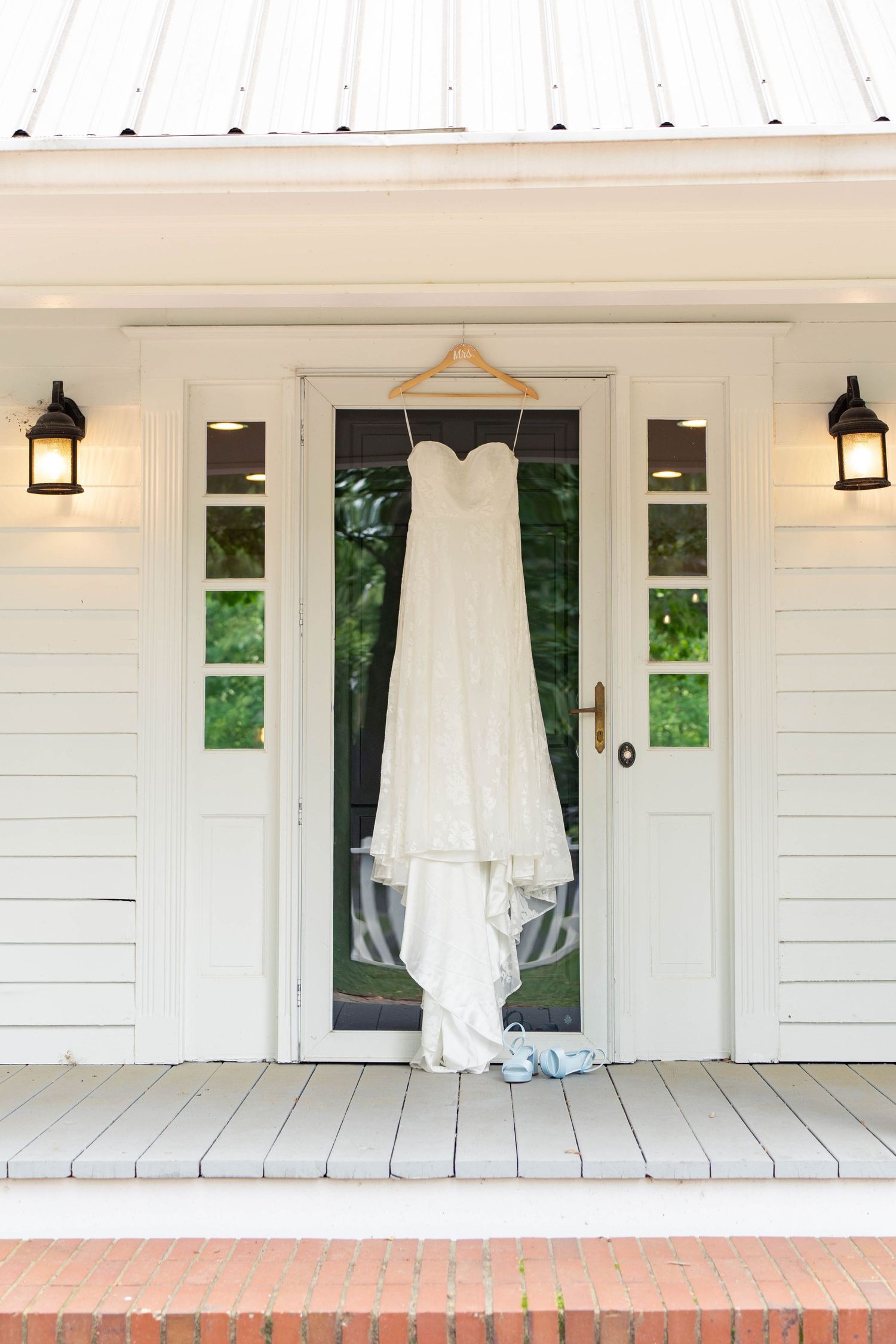 White strapless wedding dress hanging on door of charming white farmhouse porch.