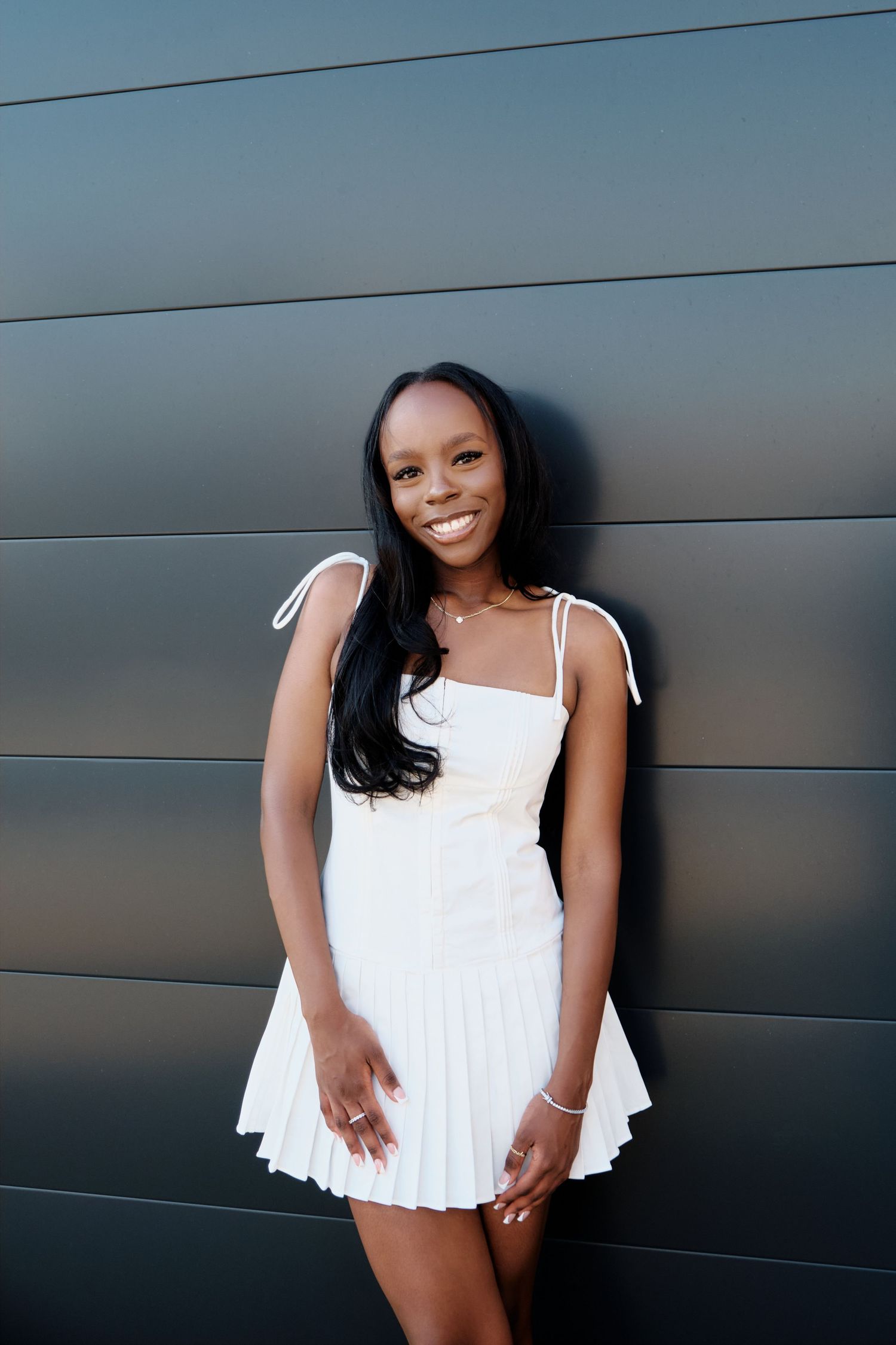 Smiling young woman in white pleated mini dress posing against a dark metallic wall.