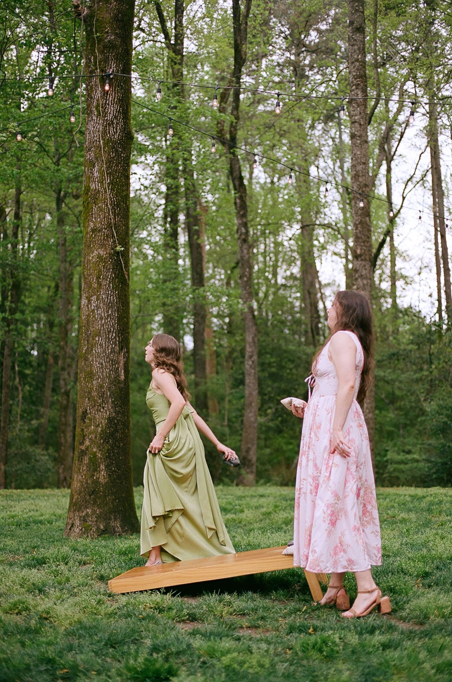 Two women in elegant dresses play cornhole in a lush green forest setting.