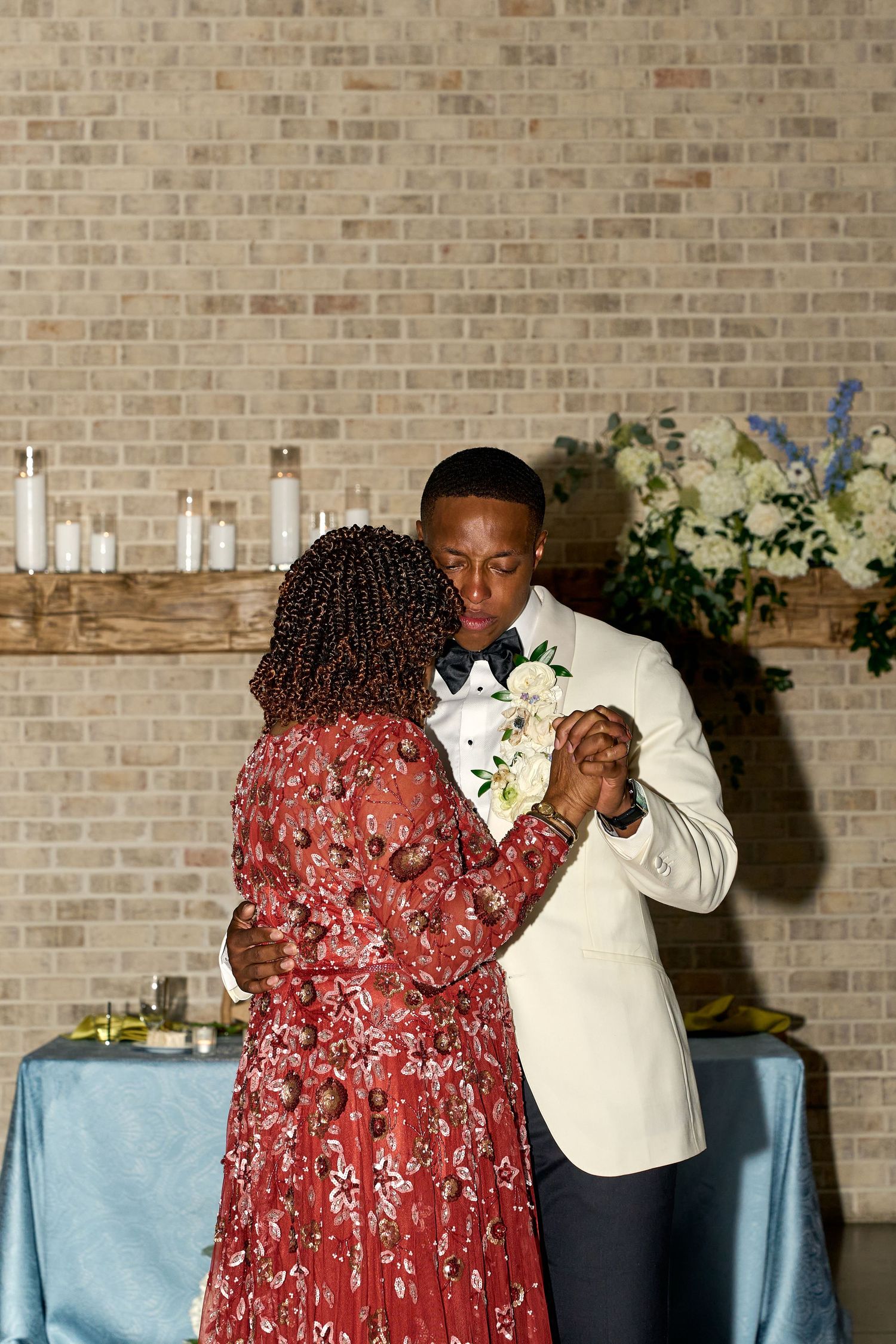 Groom in white tuxedo shares emotional mother-son dance at wedding reception before brick wall backdrop.