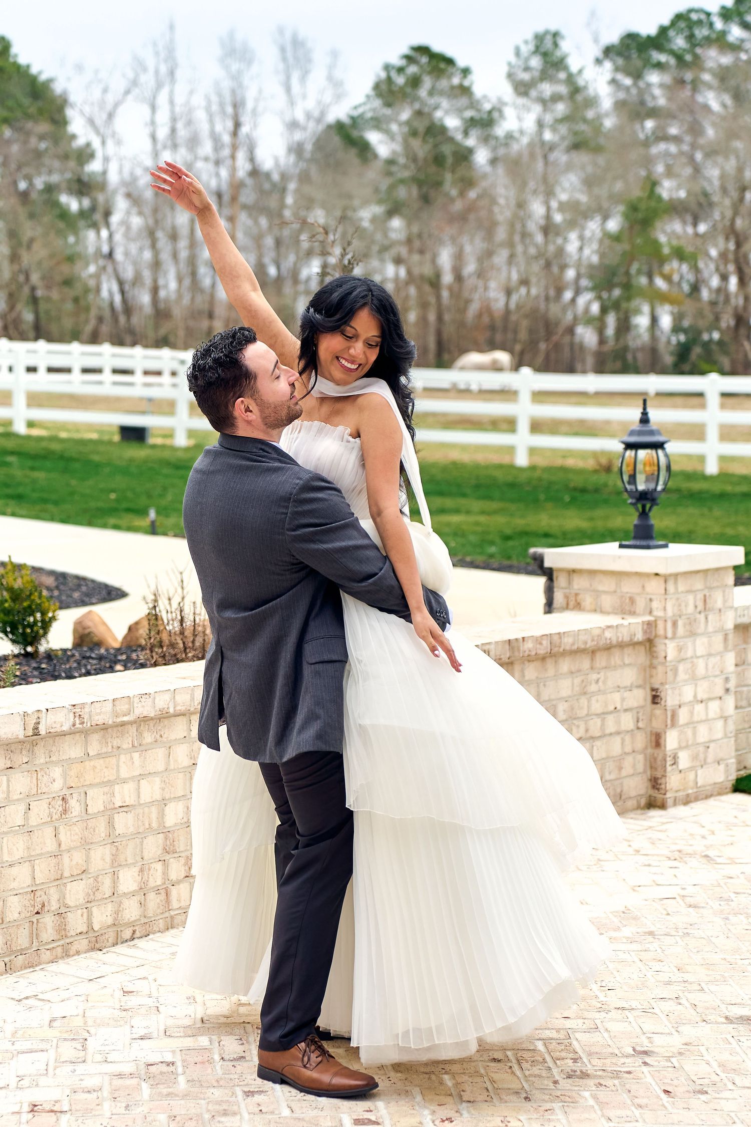 A romantic wedding couple shares a joyful moment on a paved patio with white fence and pine trees in background.