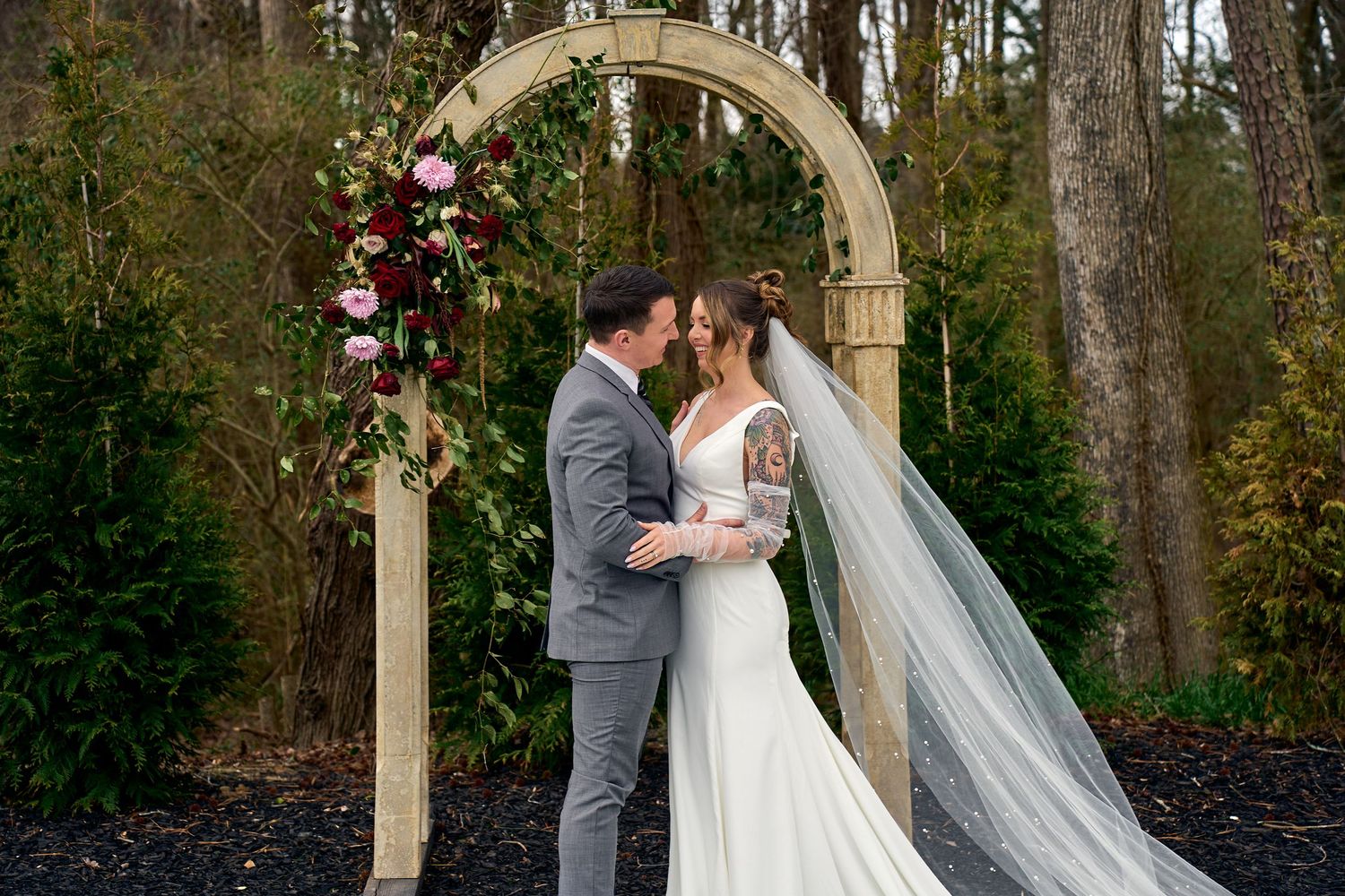 A romantic outdoor wedding ceremony under a decorated floral arch in a wooded setting.