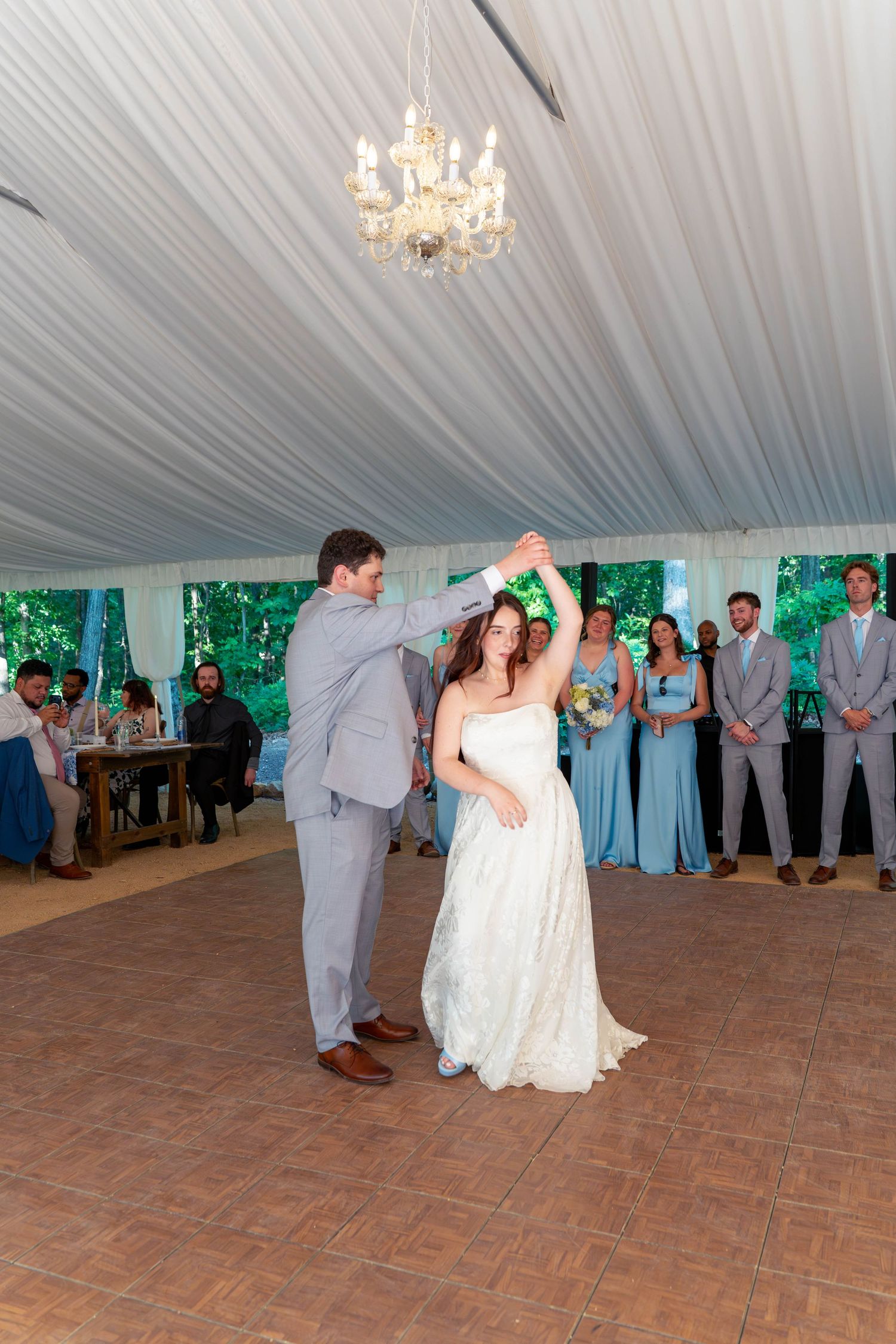 Bride and groom share first dance under chandelier as wedding party watches in white tent.