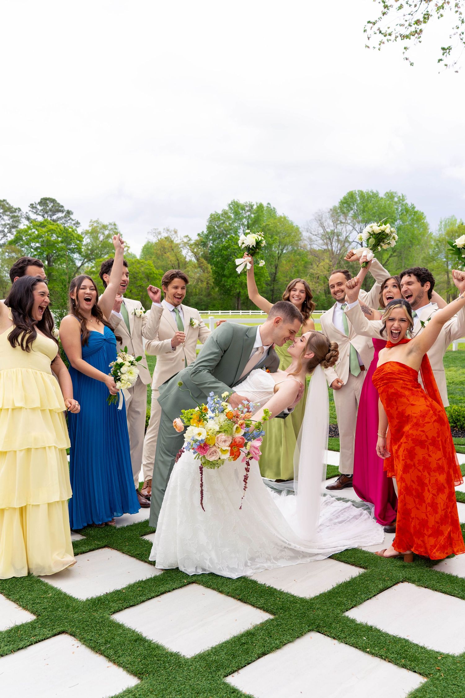 Wedding party cheers as groom dips bride for a kiss on a checkered grass aisle at outdoor ceremony.