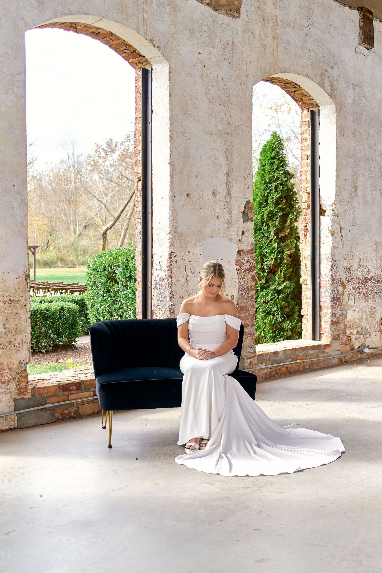 Bride in off-shoulder white gown sits gracefully on velvet sofa in rustic ruins. North Carolina Wedding Photographer.