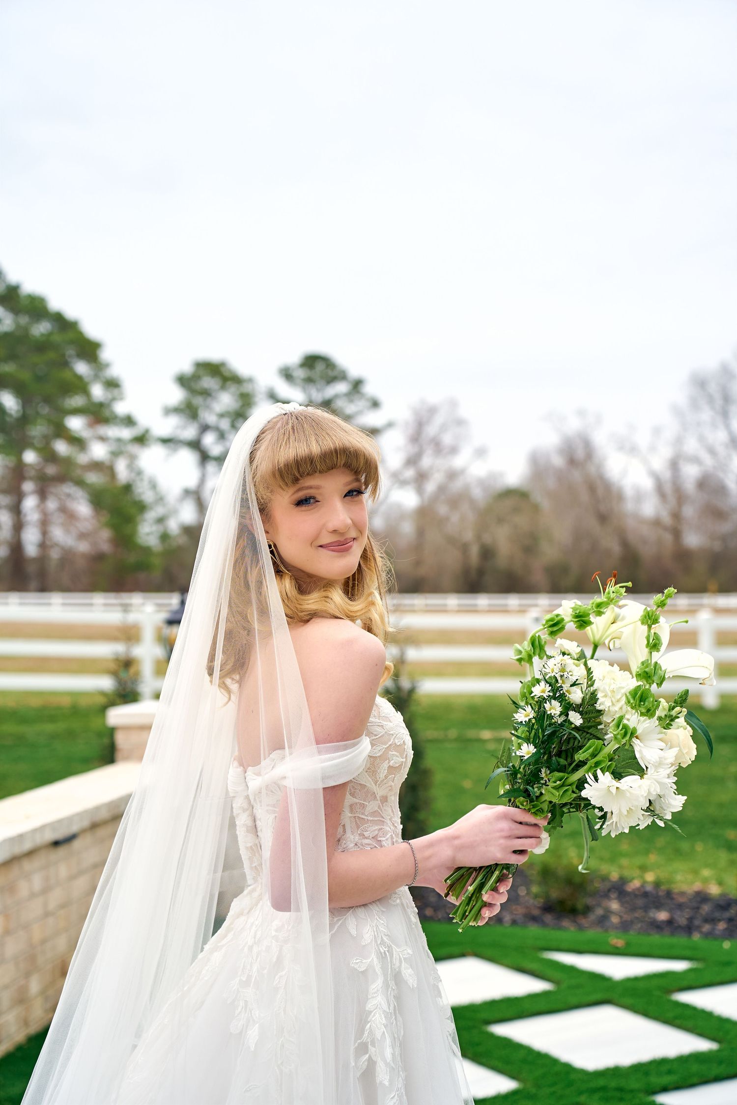 Bride in white lace wedding dress holds elegant bouquet of white flowers in manicured garden setting.