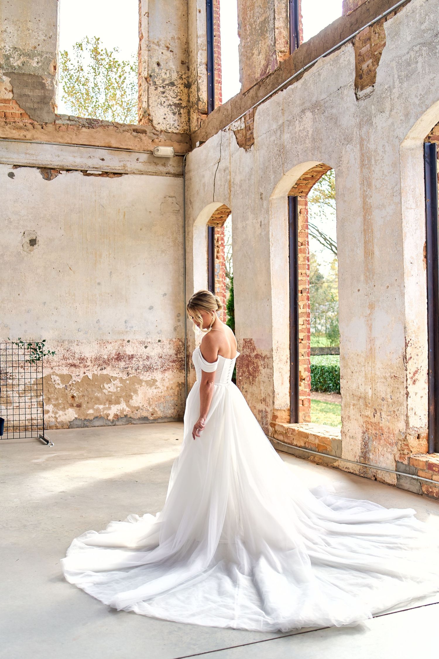A flowing white wedding gown with long train photographed in a rustic abandoned building with arched windows and weathered walls.