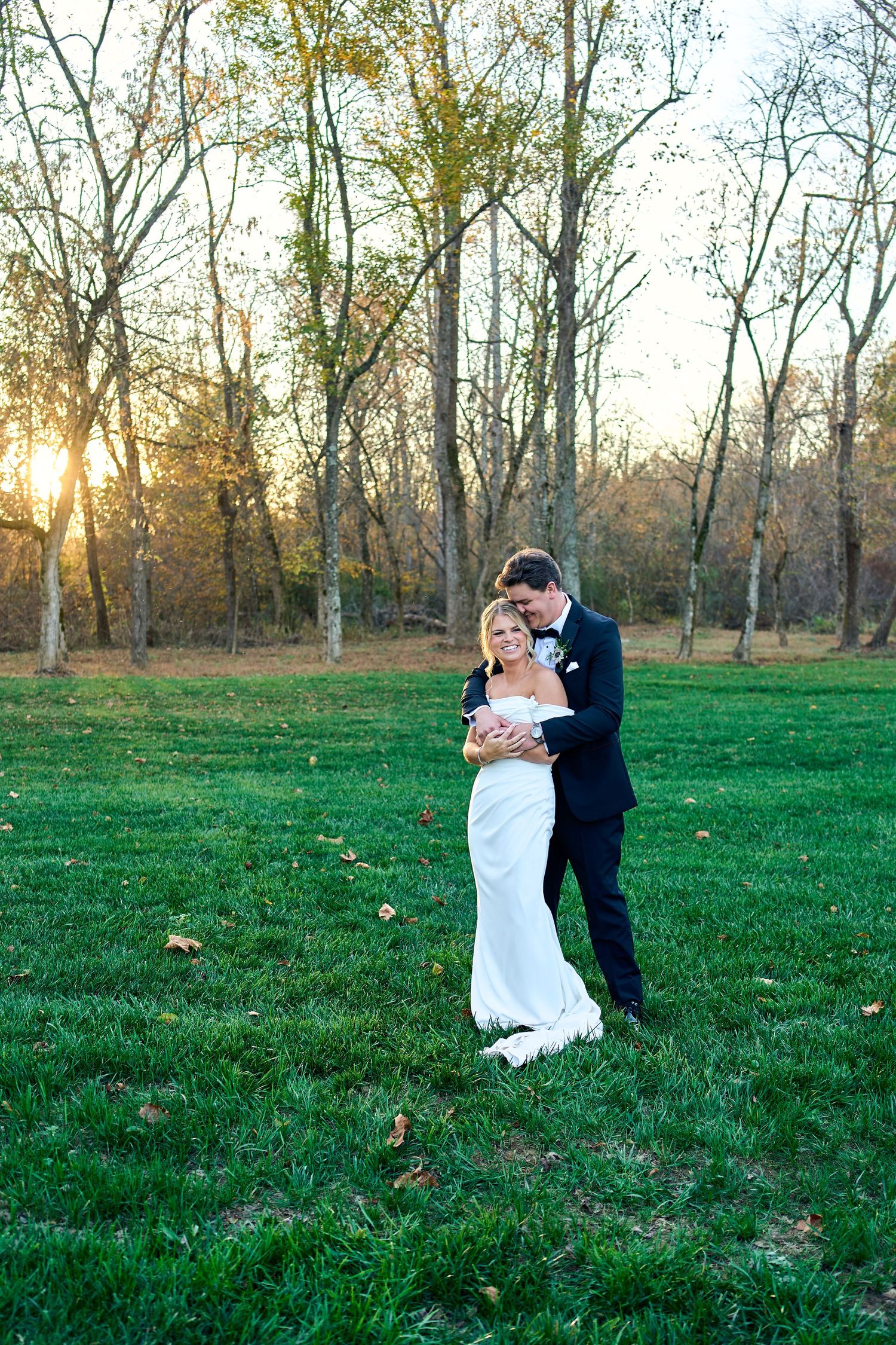 Wedding couple embraces in a park at sunset with bare trees and green grass in the background.