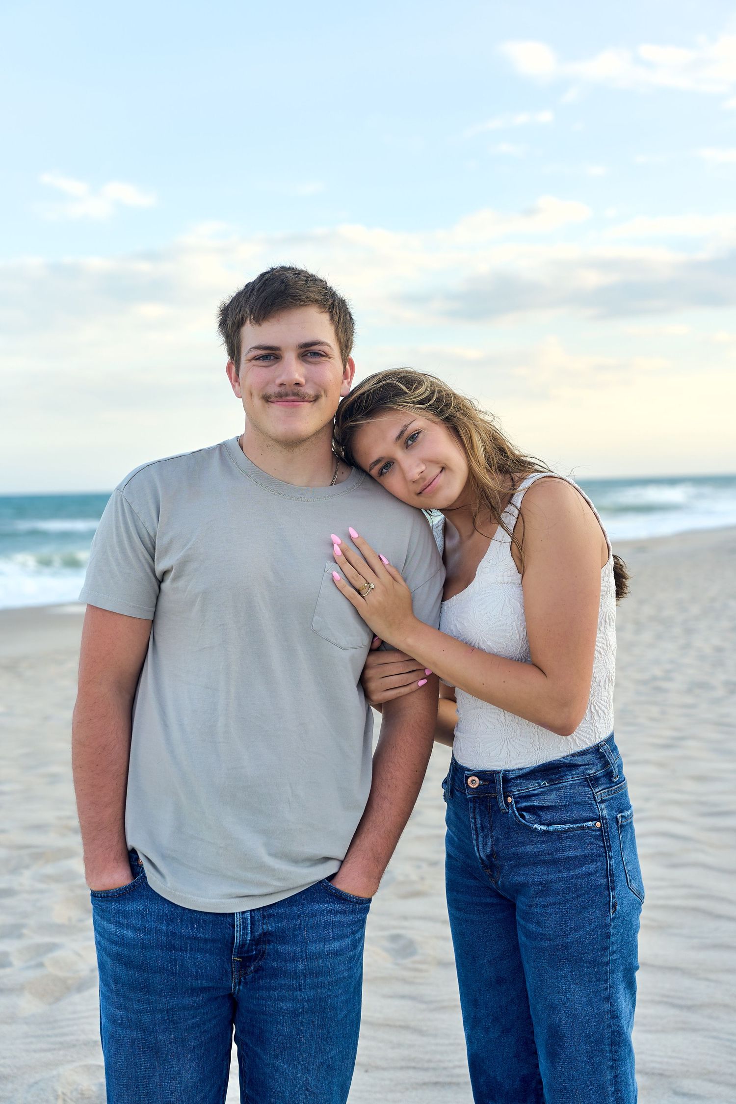 A couple poses together affectionately on a beach at sunset wearing casual jeans and t-shirts.