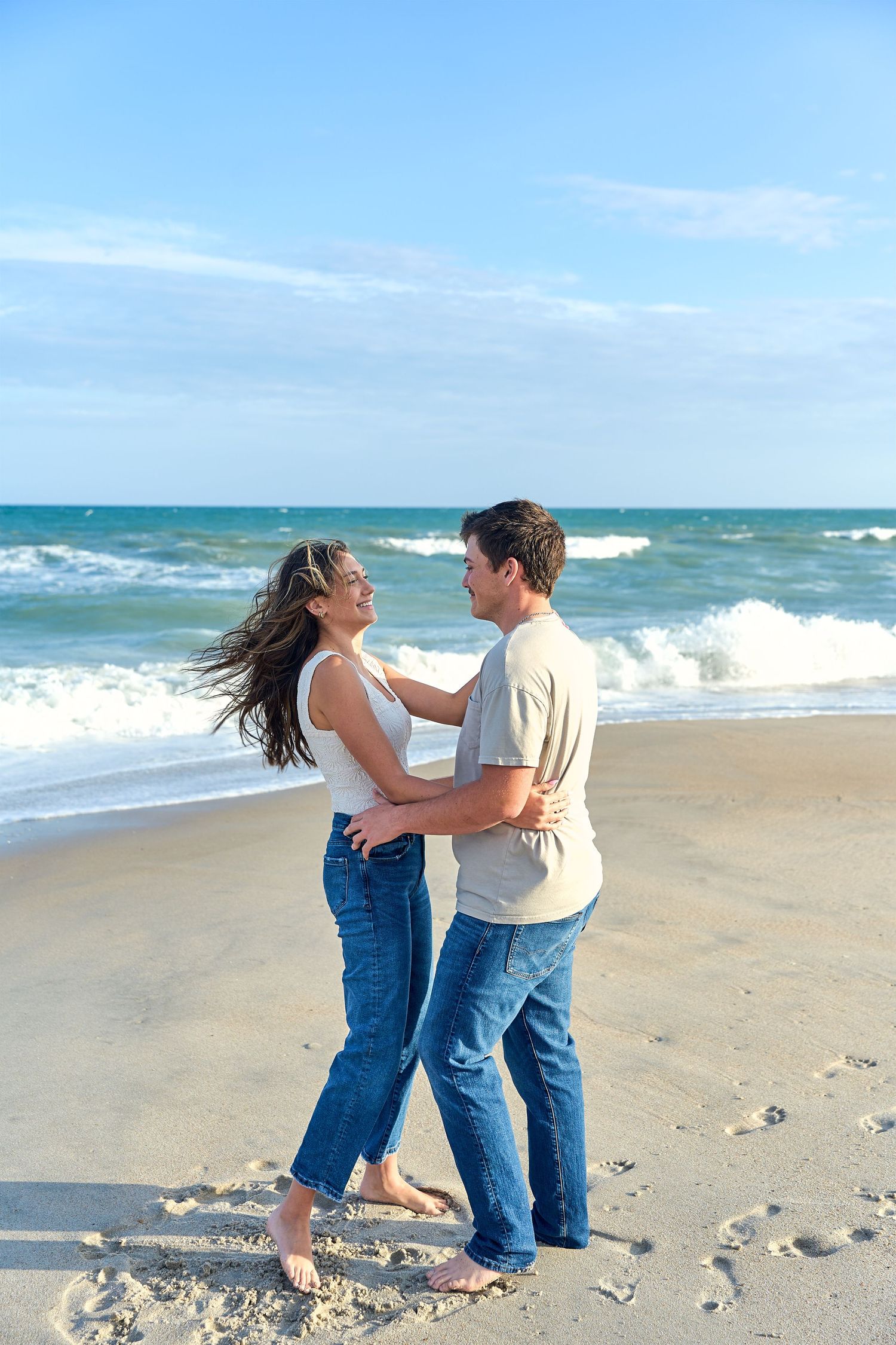 Two people embrace and dance together on a sunny beach with turquoise waves crashing in the background.