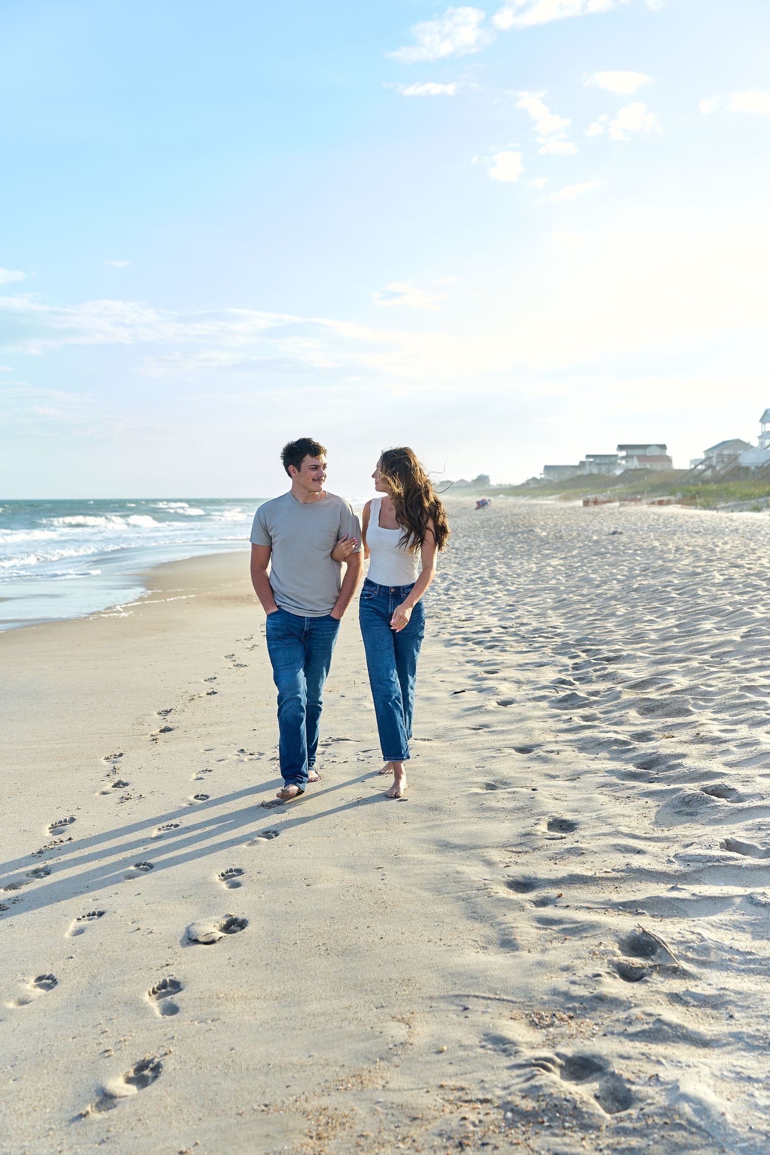 Couple walking together on a sandy beach with ocean waves and beach houses in the background.