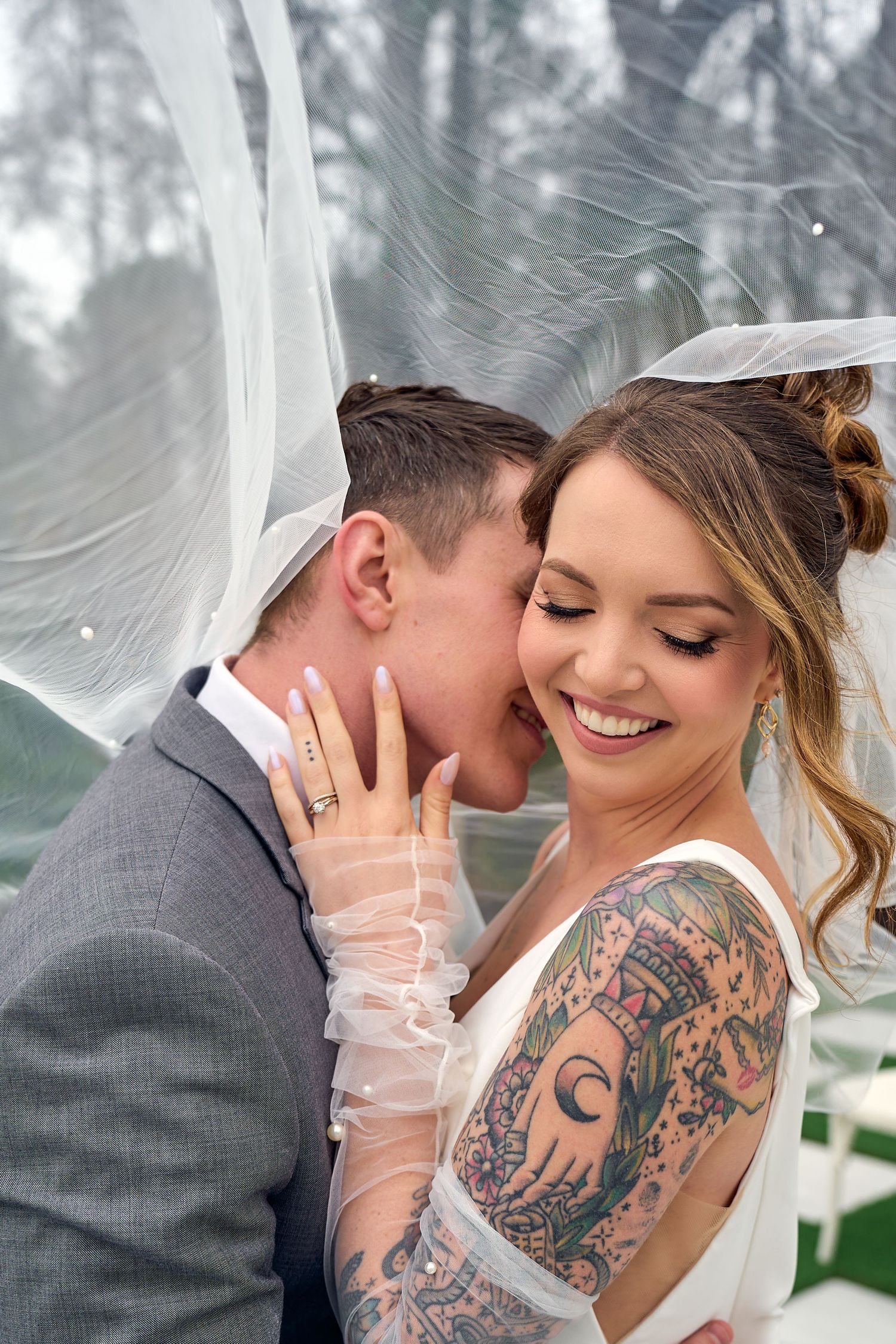 Intimate wedding moment shows couple embracing under a veil, with bride displaying elegant tattoo sleeve and sheer gloves.