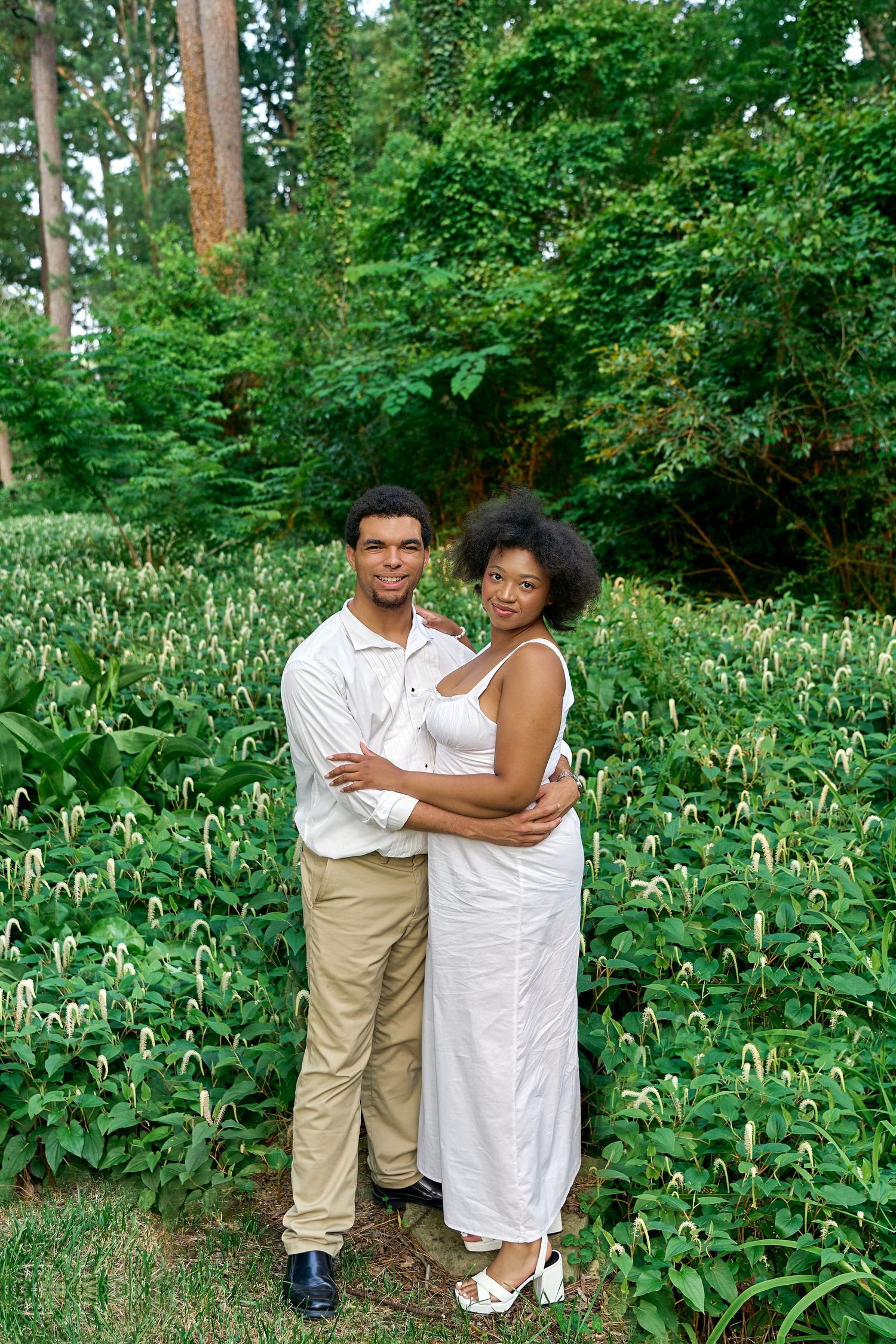 A romantic couple embraces in a lush garden setting with tall trees and greenery.