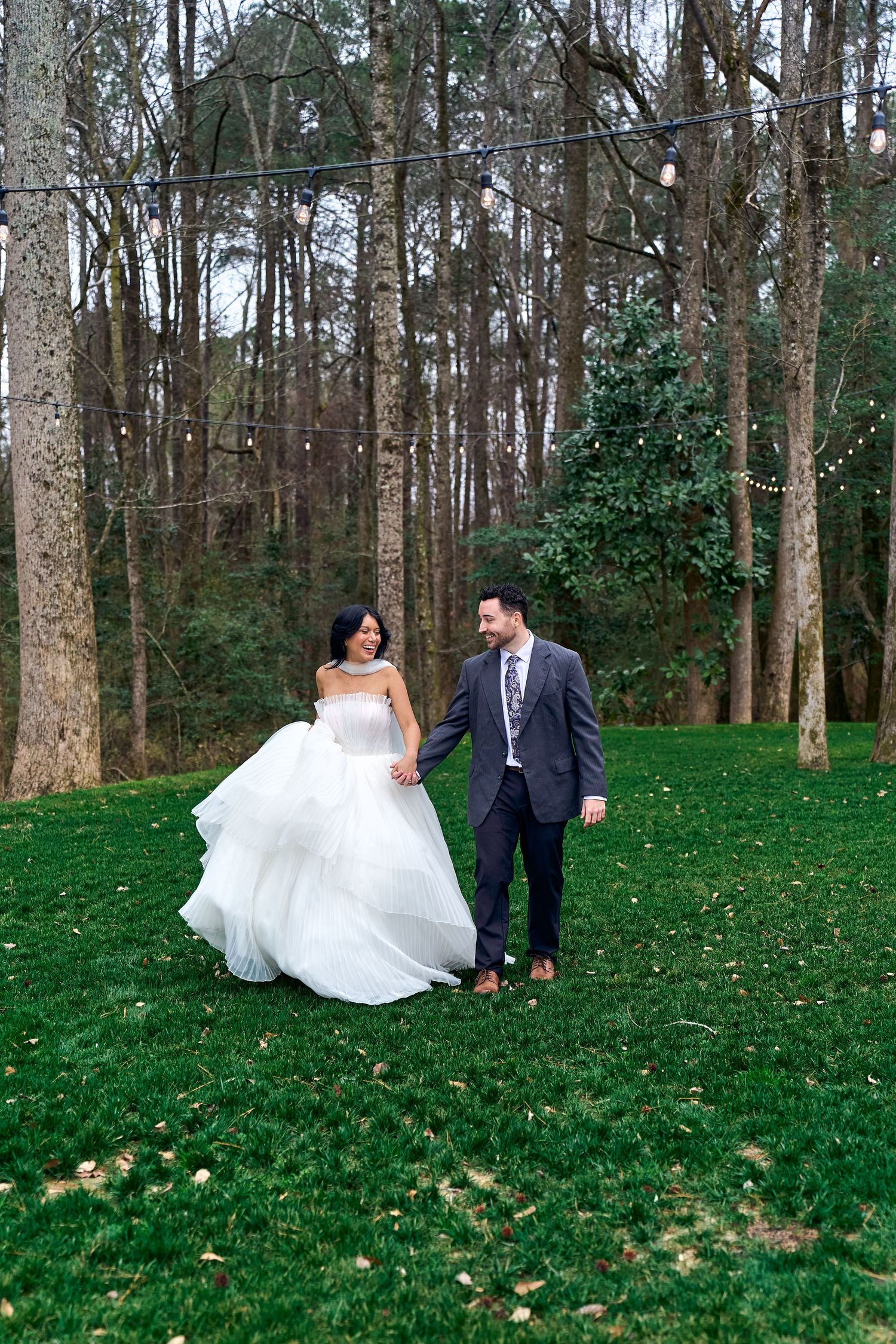 Amidst a wooded setting, two people celebrate their wedding day on a lush green lawn surrounded by tall bare trees.