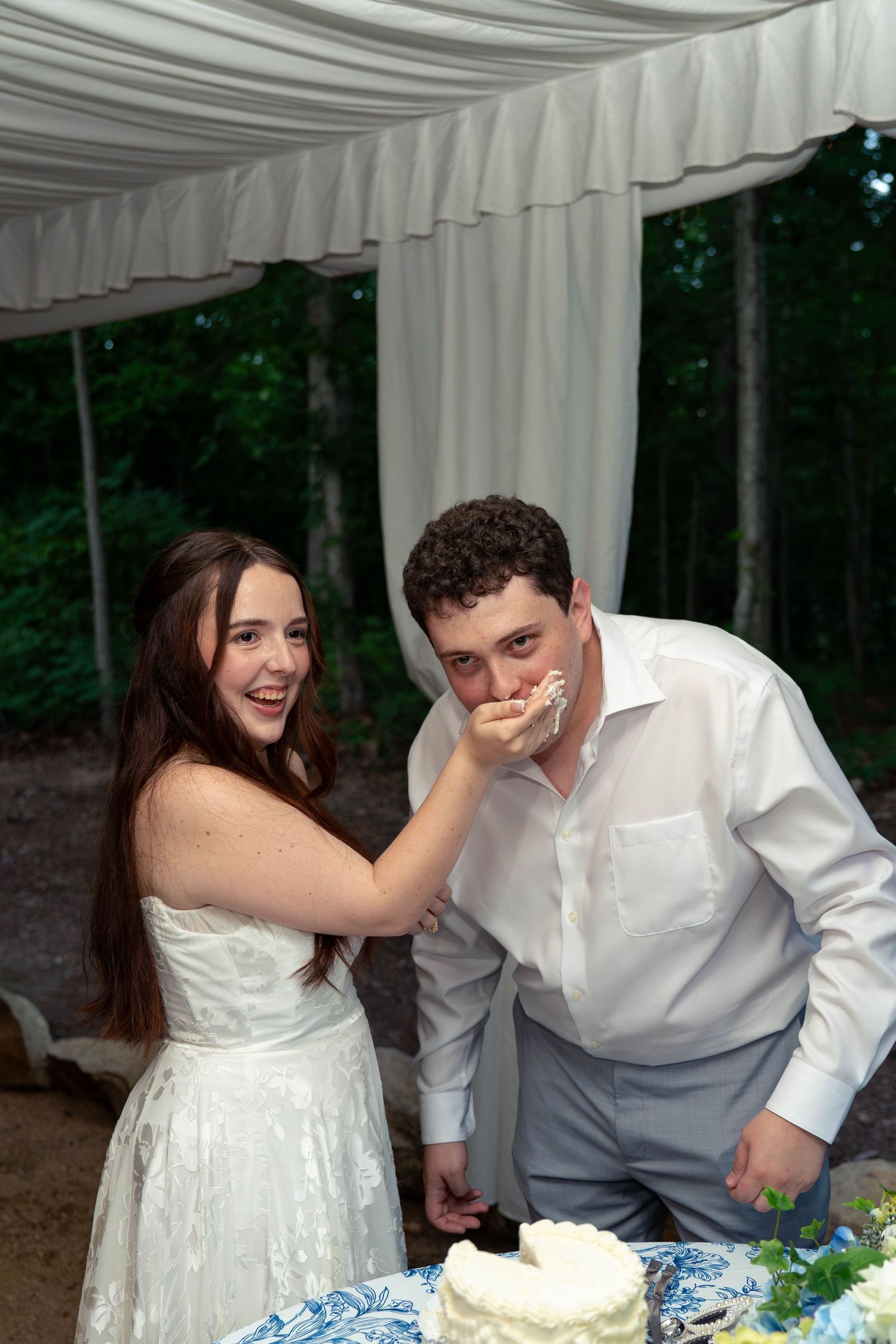 Bride feeds groom wedding cake outdoors under white tent at reception.