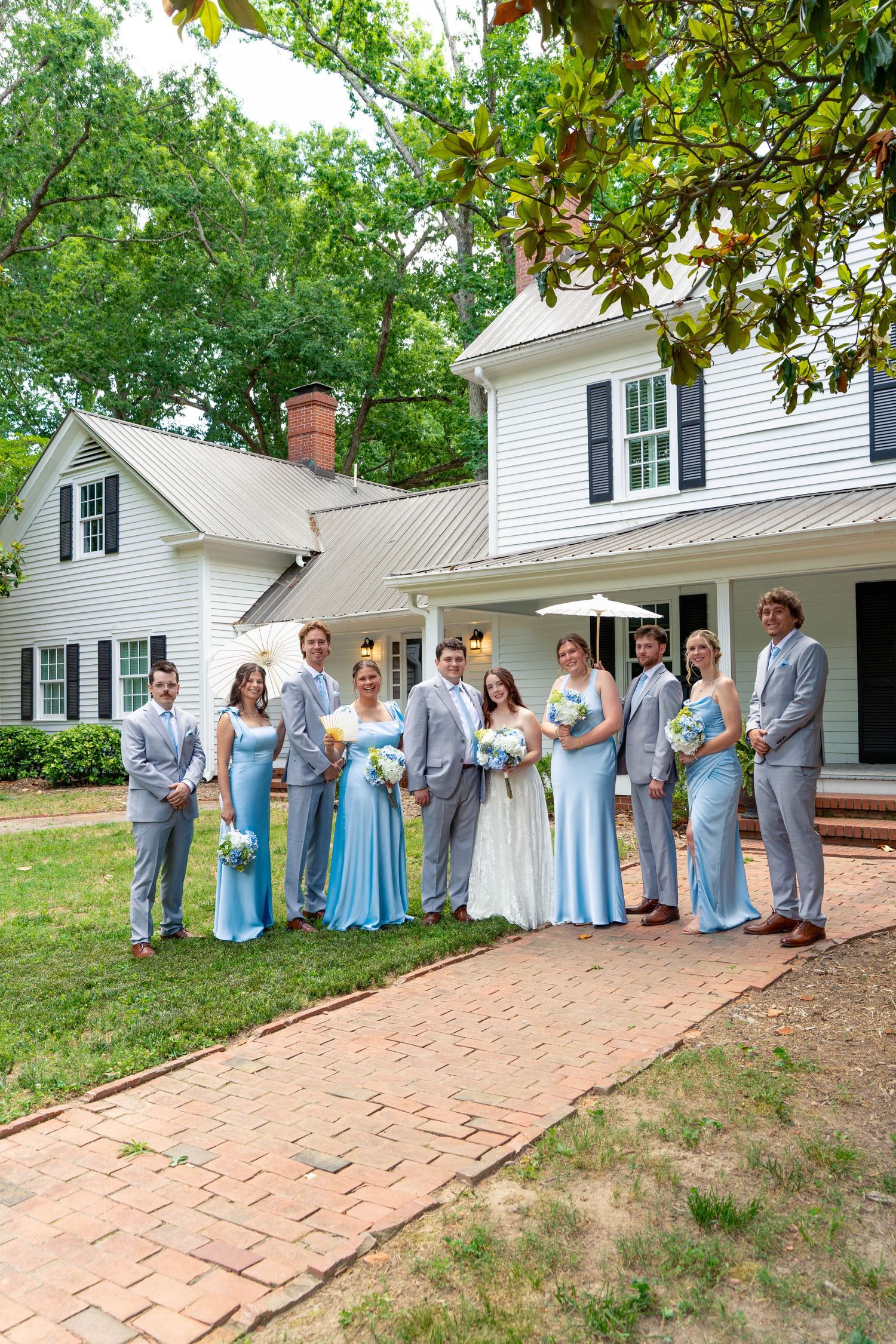 Wedding party in light blue dresses and gray suits poses outside white colonial home.