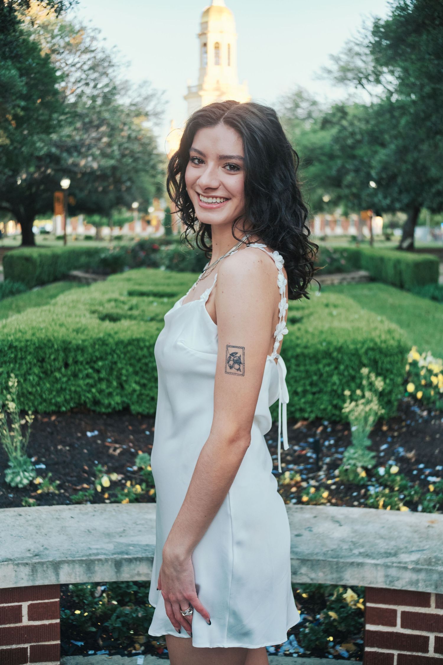 Smiling young woman in white slip dress with tattoo posing in a garden with a clock tower behind her.