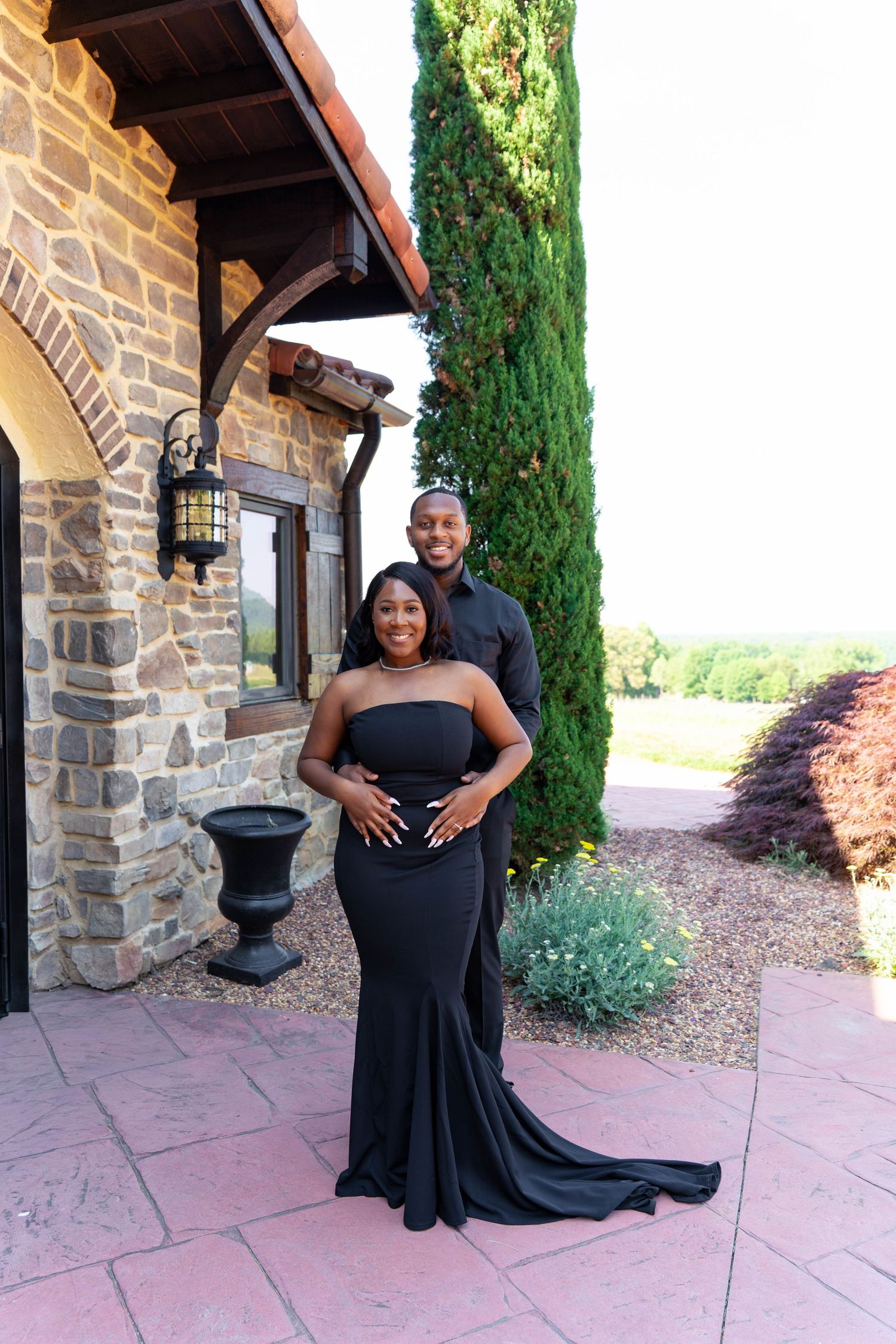 Couple in matching black formal attire posing together outside a rustic stone building with cypress trees.