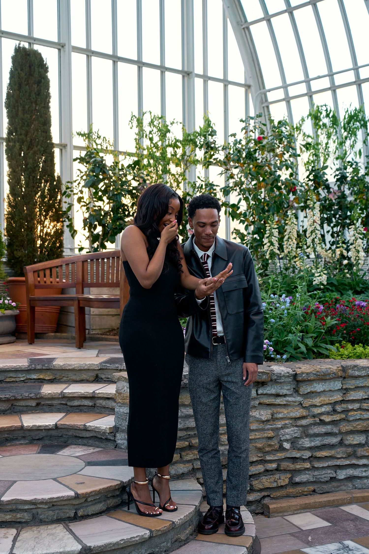 Couple admiring an engagement ring inside a sunlit botanical conservatory surrounded by lush greenery.