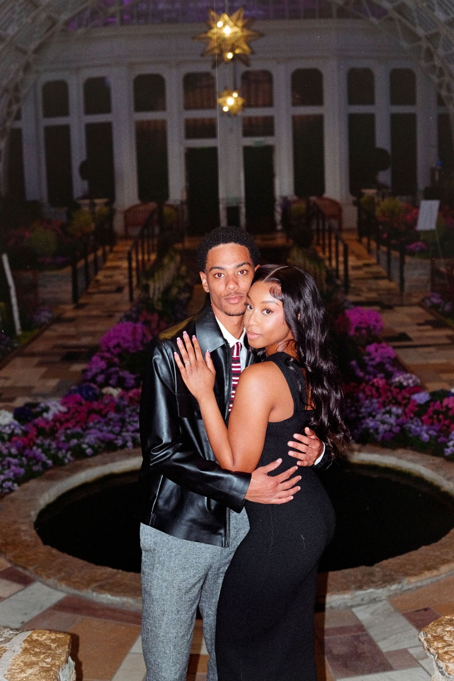 Couple embraces in stunning conservatory surrounded by vibrant purple flowers at night.