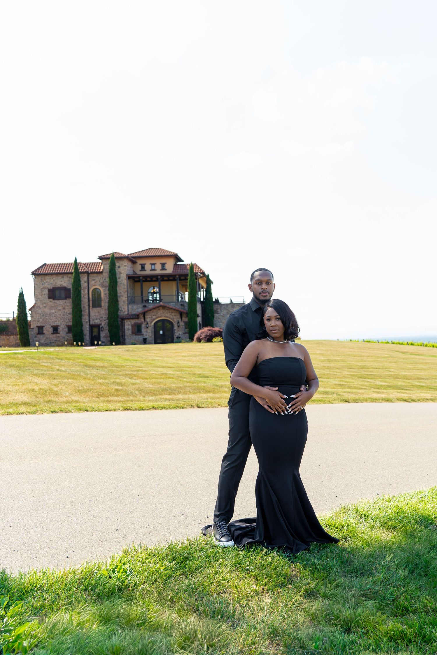 Couple posing intimately in black formal wear before a grand Tuscan-style mansion on a sunny day.