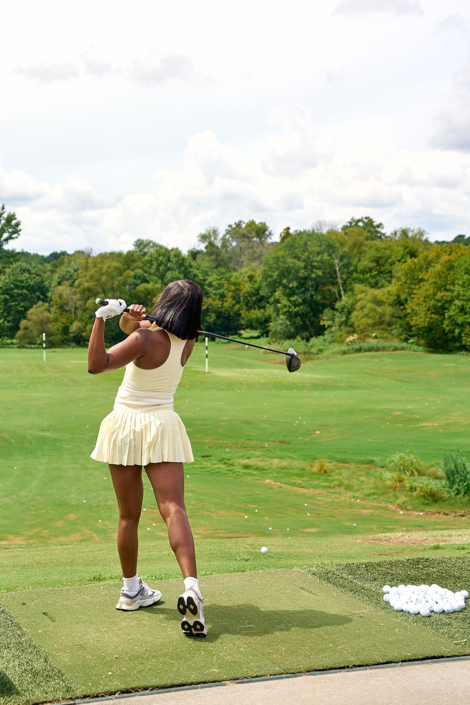 Woman in yellow tennis skirt completes golf swing follow-through at lush green driving range.