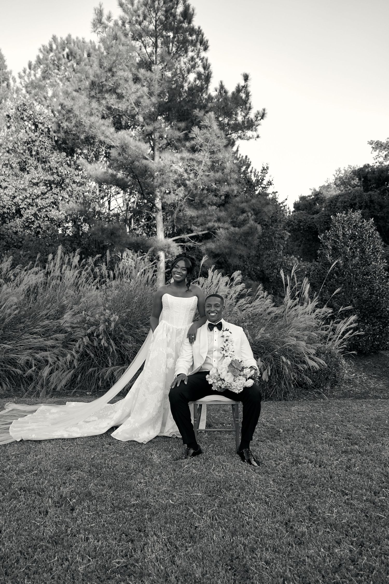 Black and white photo of laughing bride standing beside seated groom holding bouquet in garden setting.
