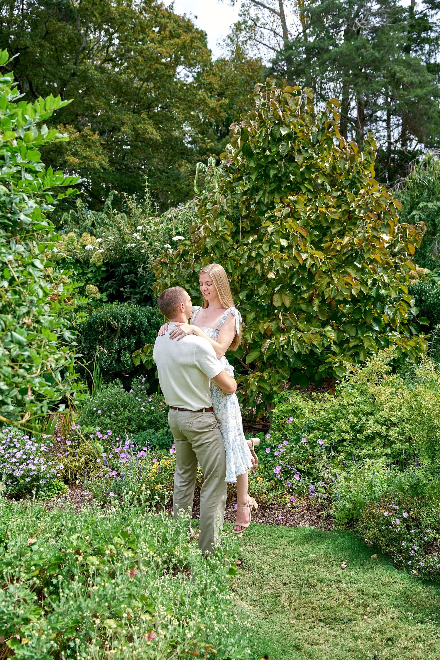 Couple dancing and laughing together in a lush green garden surrounded by colorful flowering plants.