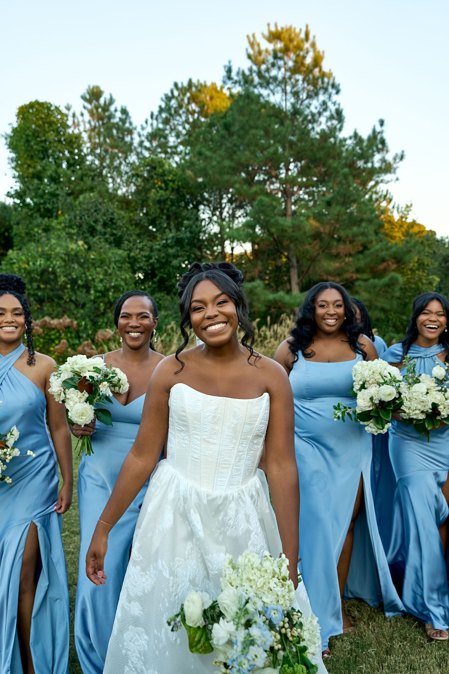 Smiling bride in white gown leads bridesmaids in blue satin dresses holding white floral bouquets outdoors.