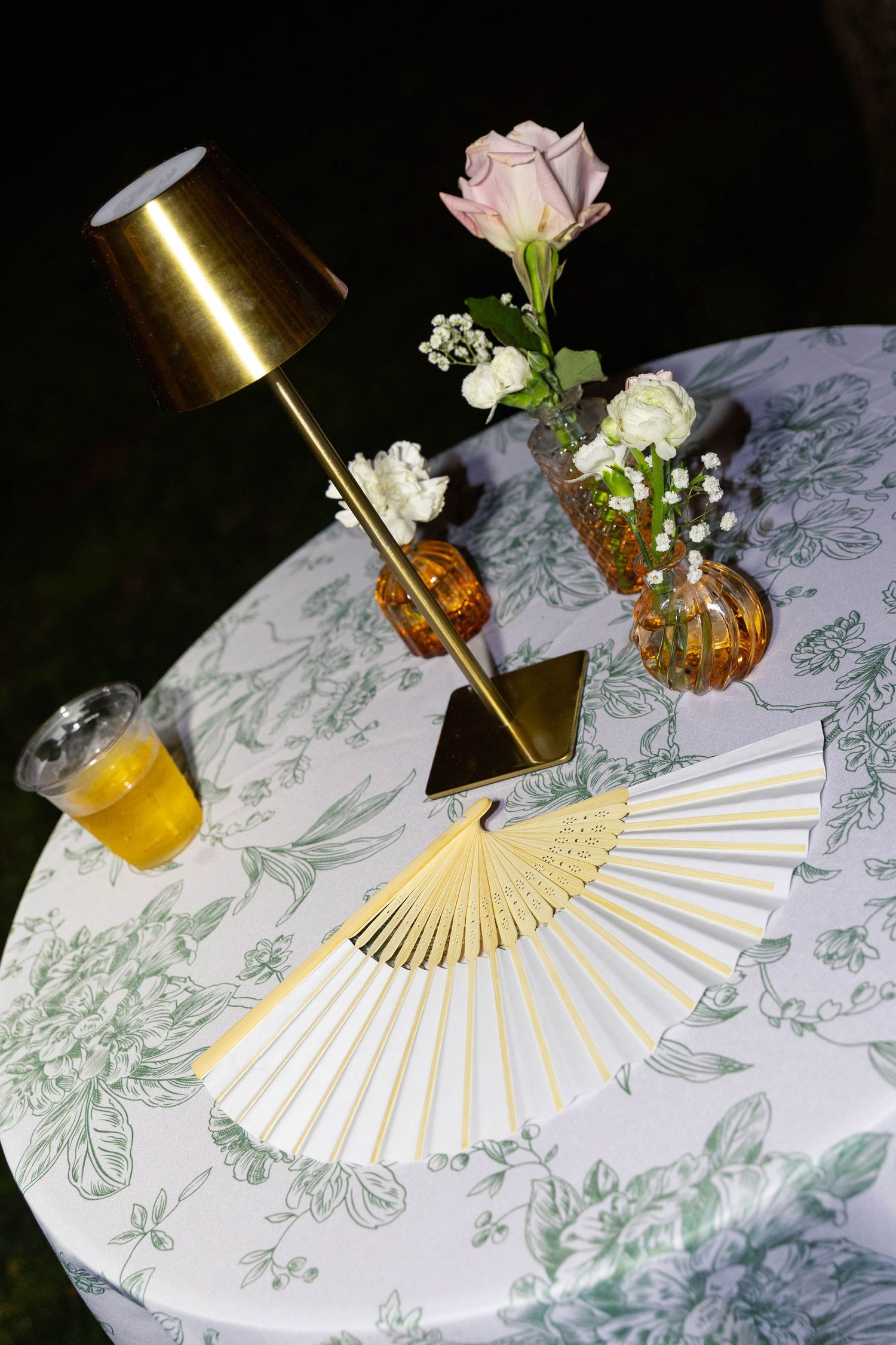 Wedding cocktail table with gold fan favors, floral arrangements, and a gold lamp at night.
