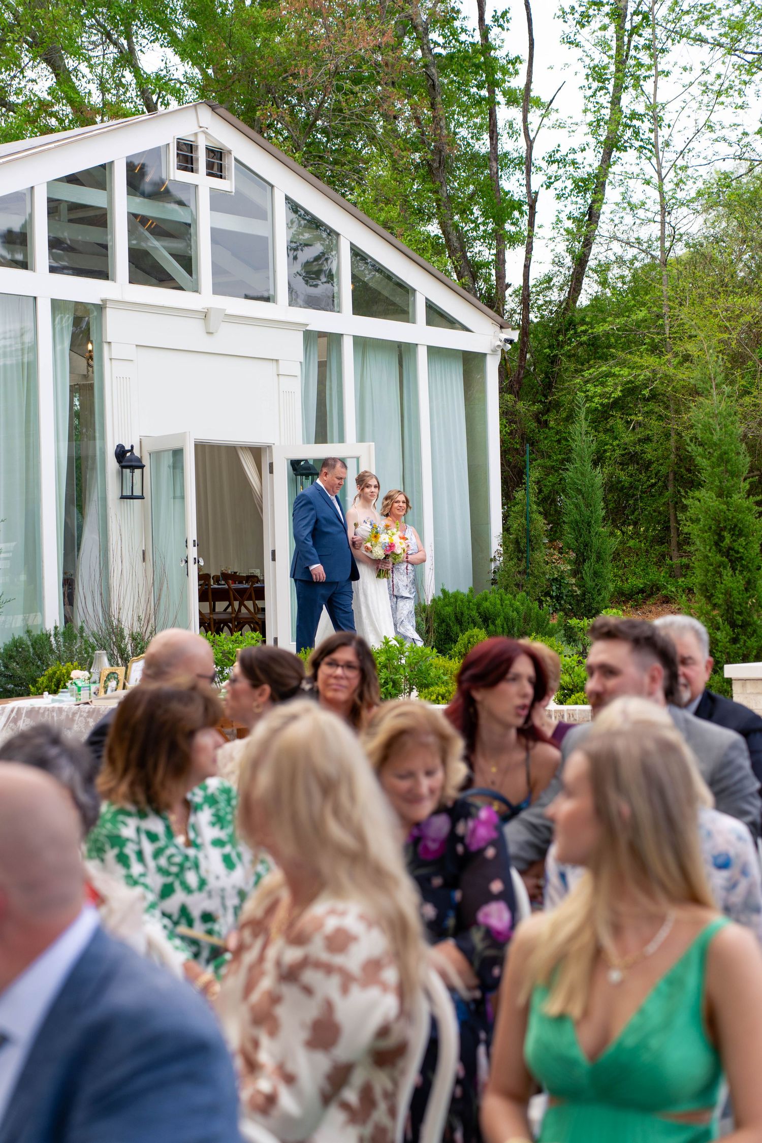 Bride and escort emerge from glass greenhouse chapel as wedding guests stand in anticipation.
