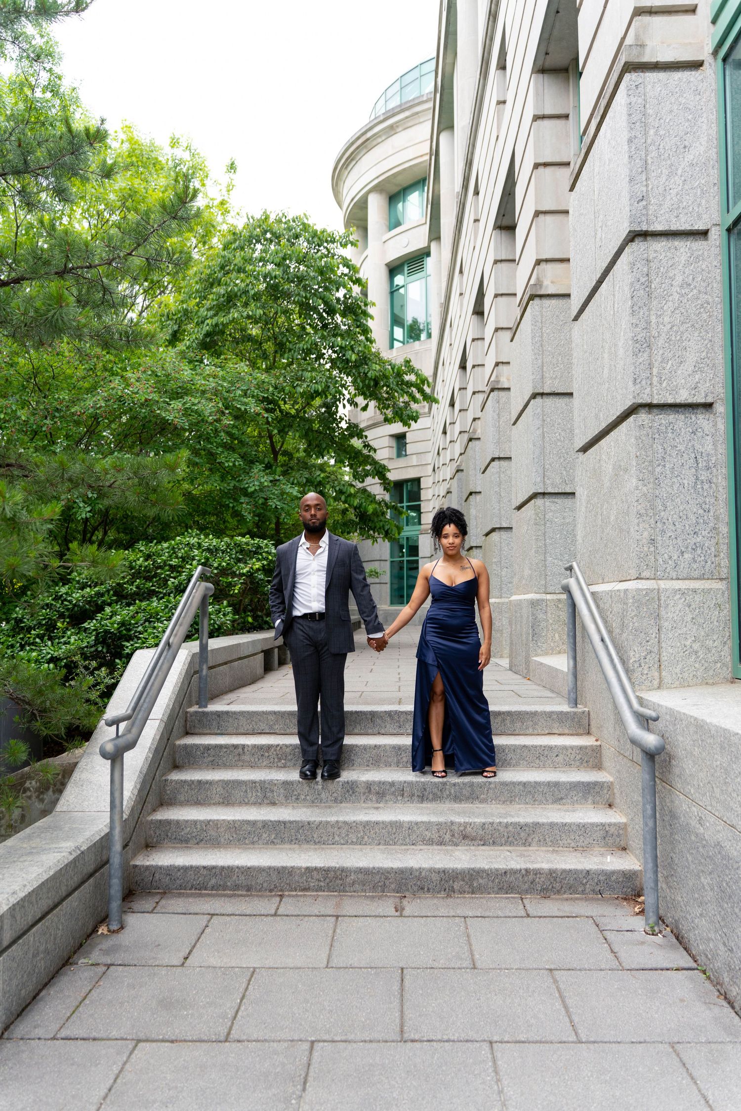 An elegant couple in formal wear holds hands while descending stone steps beside a grand building.