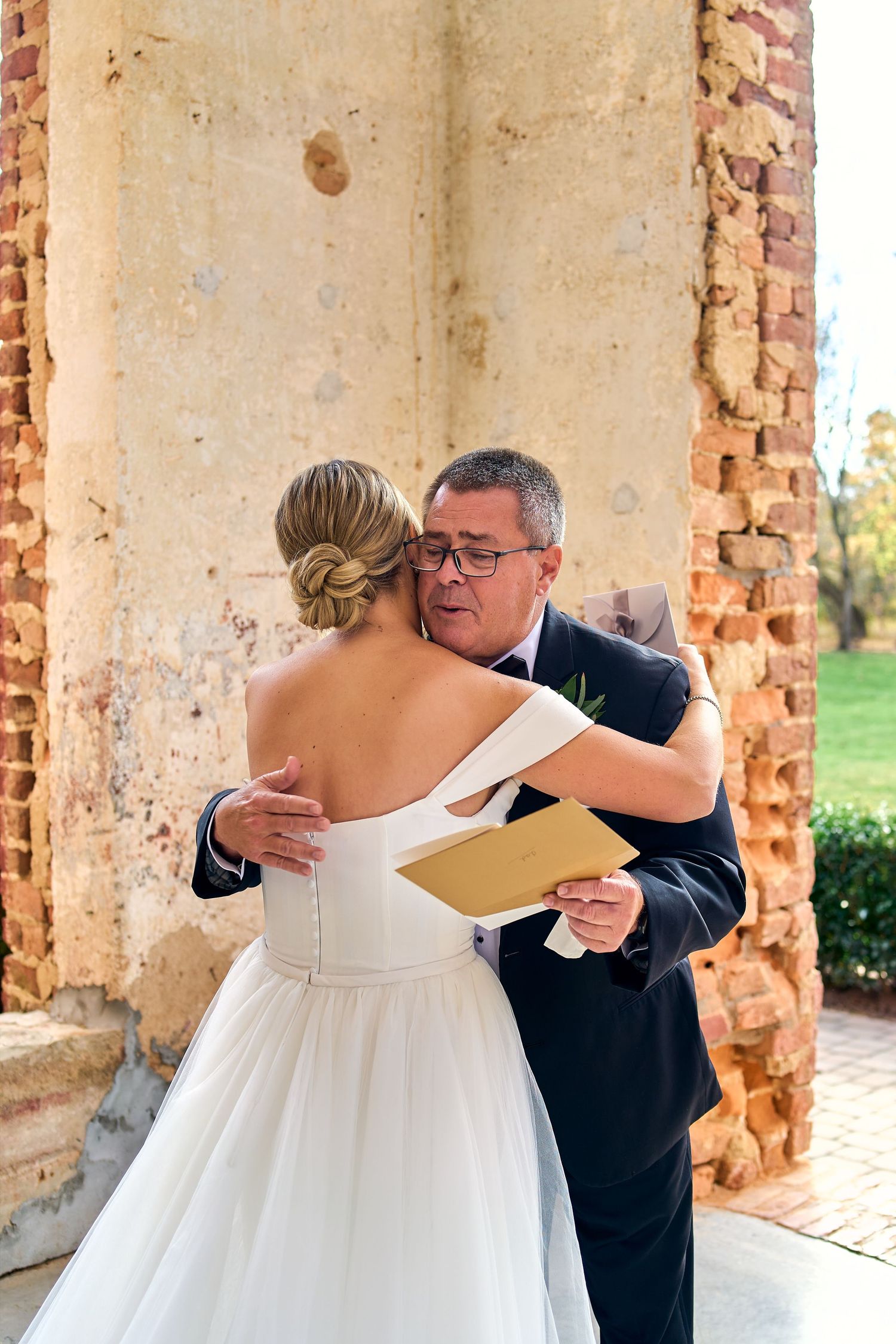 A tender wedding moment shared between a bride in a white off-shoulder gown and someone holding papers during an outdoor ceremony.