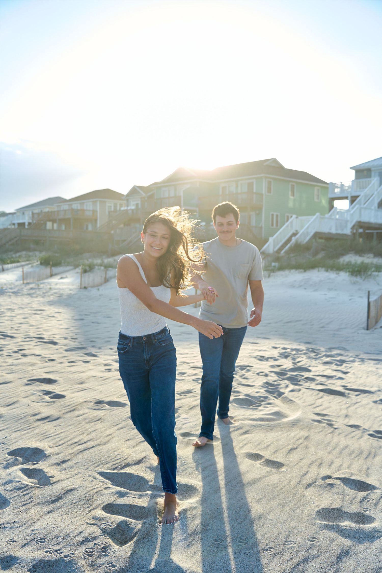 A couple runs and laughs together on a sandy beach at sunset with beach houses visible in the background.