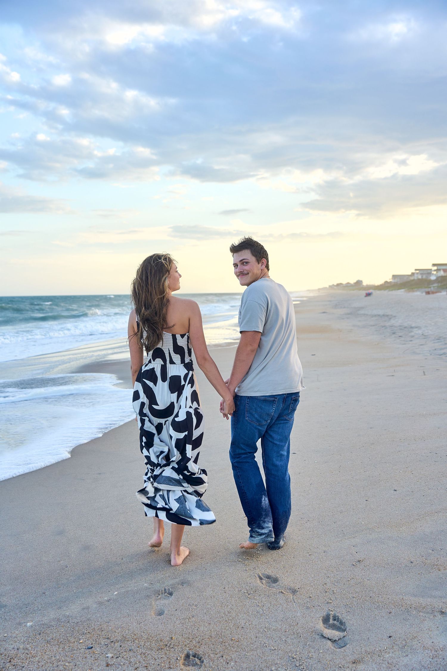 A couple walks hand in hand along a sandy beach at sunset with ocean waves and cloudy sky in the background.
