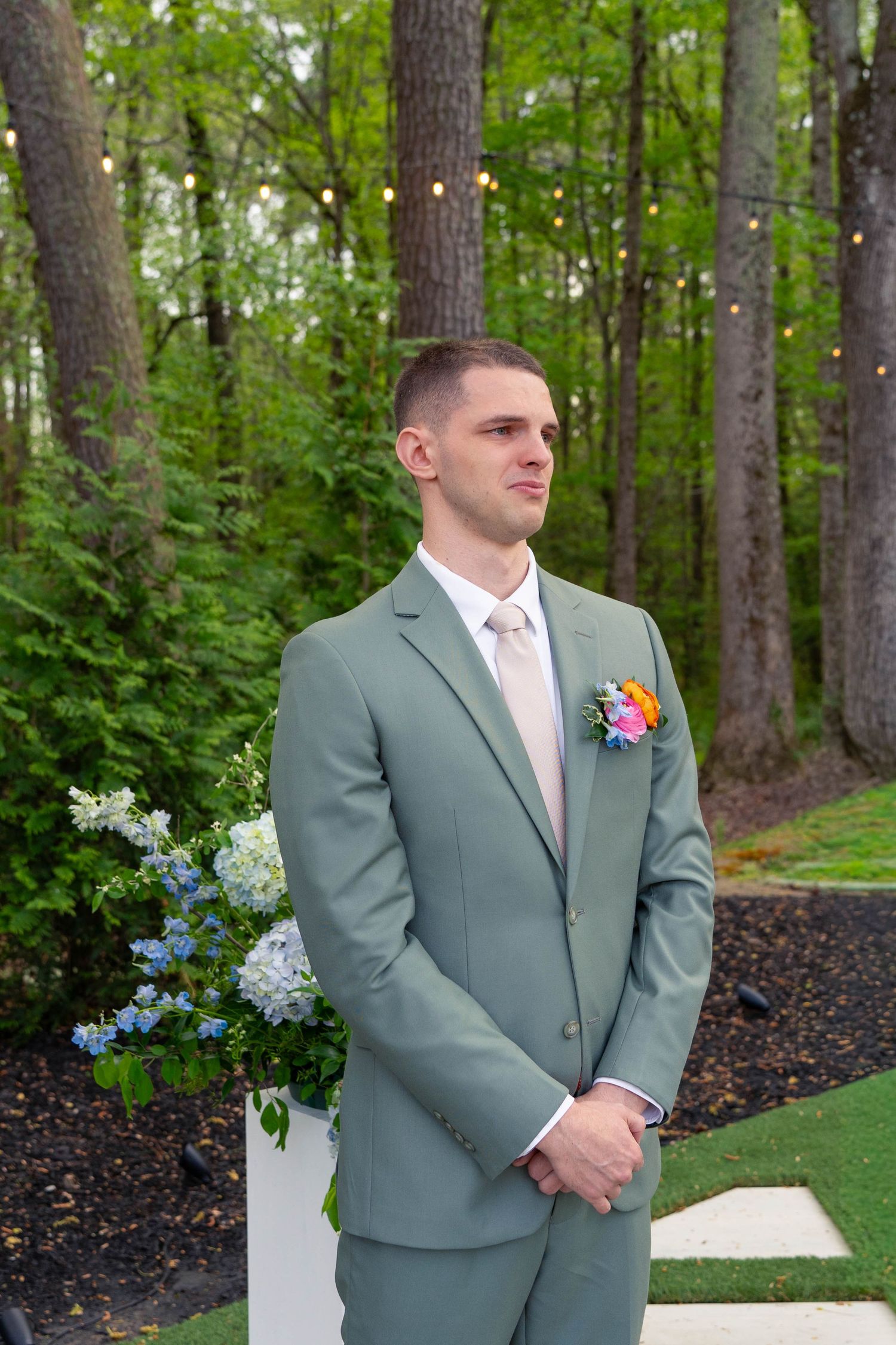 Emotional groom in sage green suit with floral boutonniere awaits bride at outdoor wedding altar.